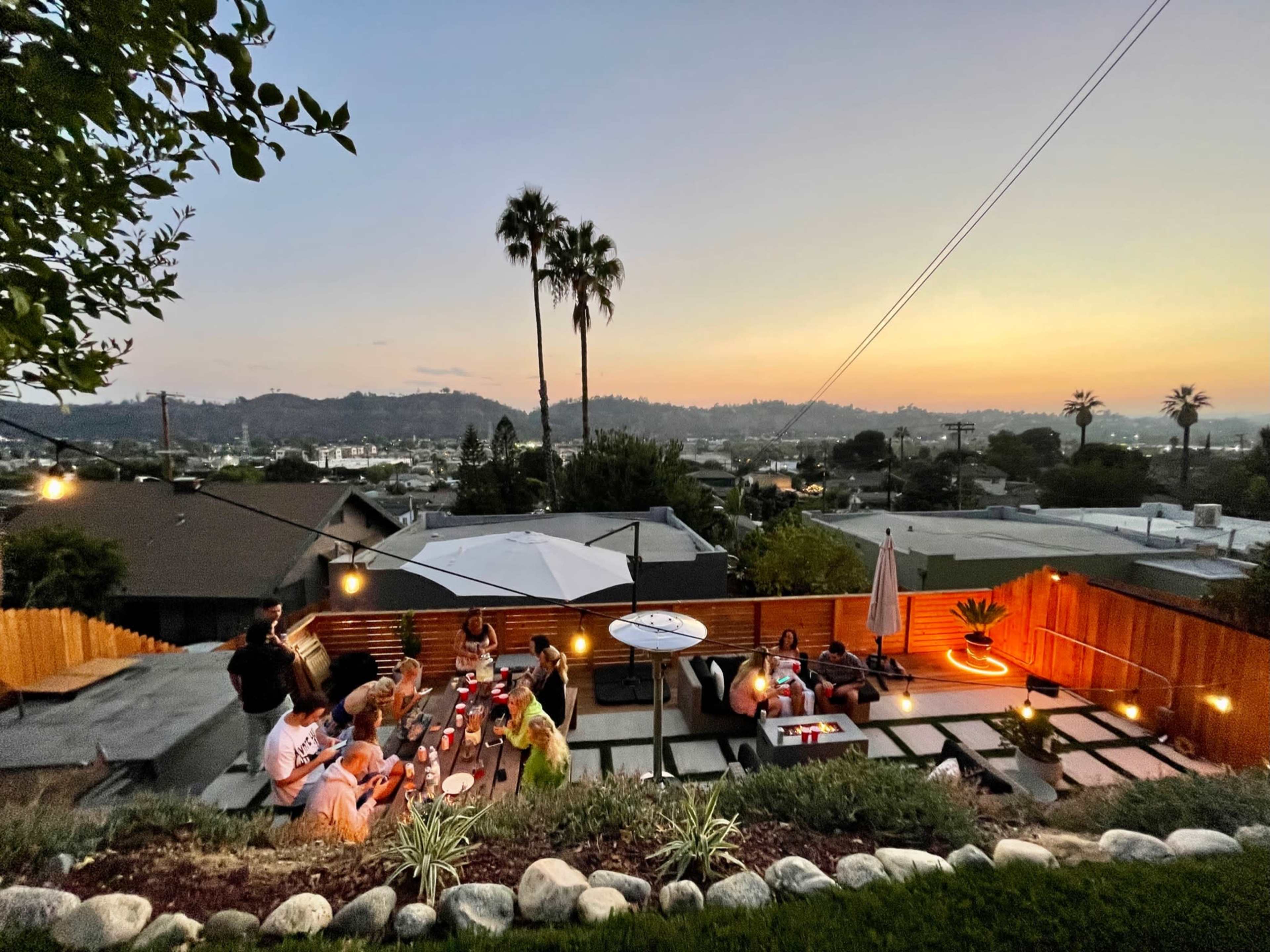 A large group of people gathers for an evening dinner on a hillside patio, with city lights and mountains visible in the background at sunset.