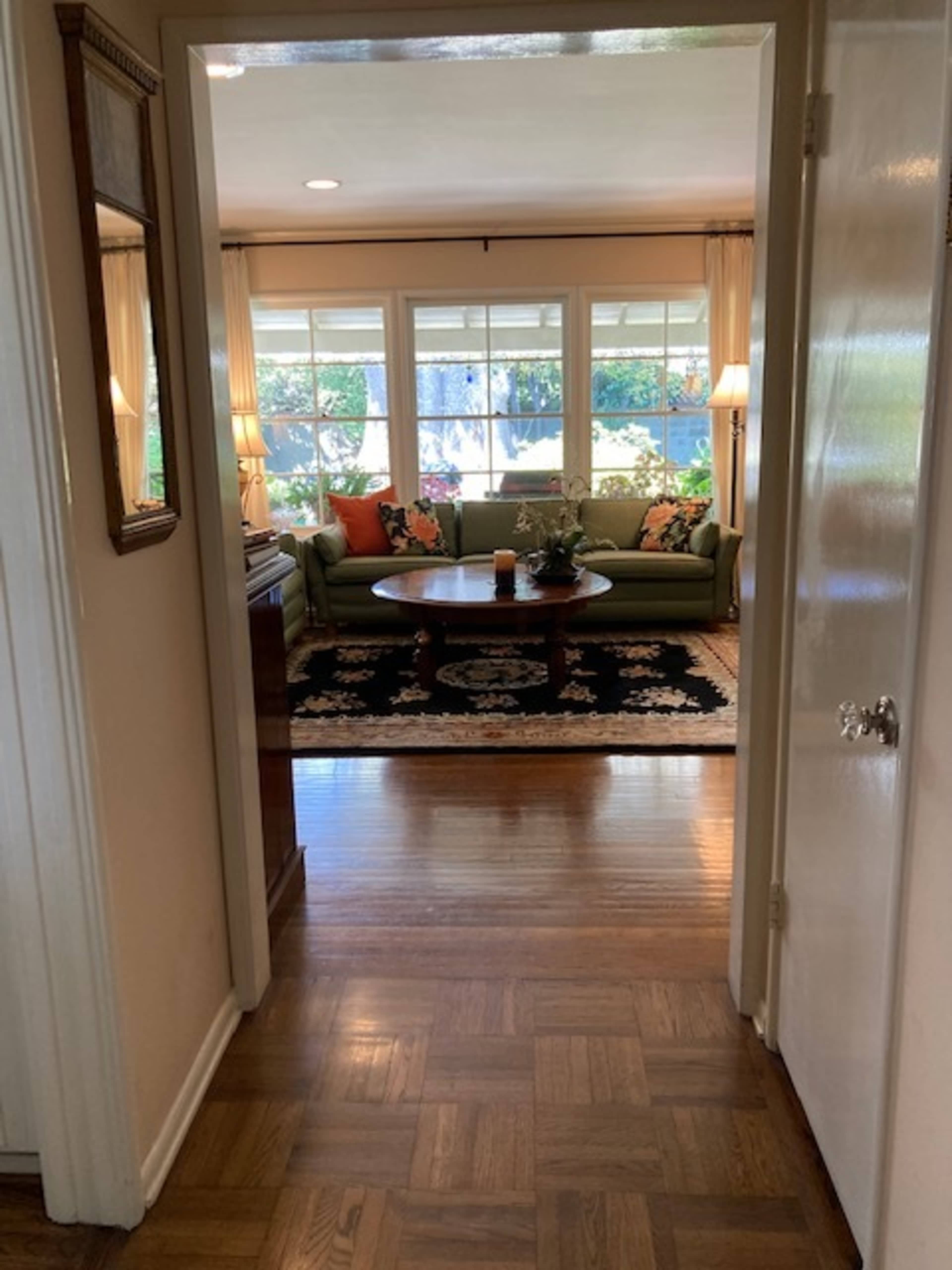 A hallway leading to a living room with a green sofa, a round coffee table, and large windows that let in natural light.