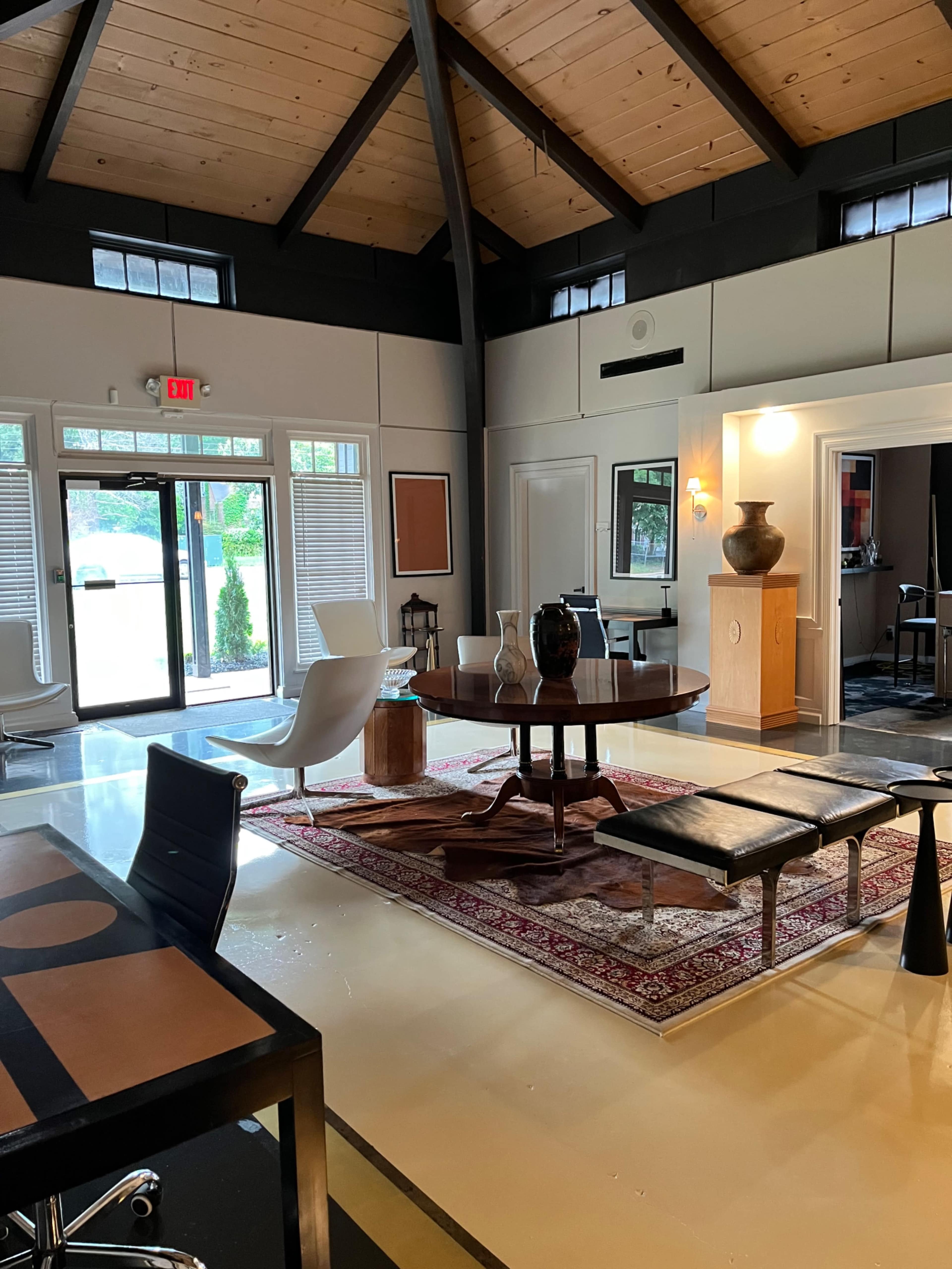 The image shows a modern interior of an office or living space featuring a round wooden table, various chairs, and decorative elements on a rug.