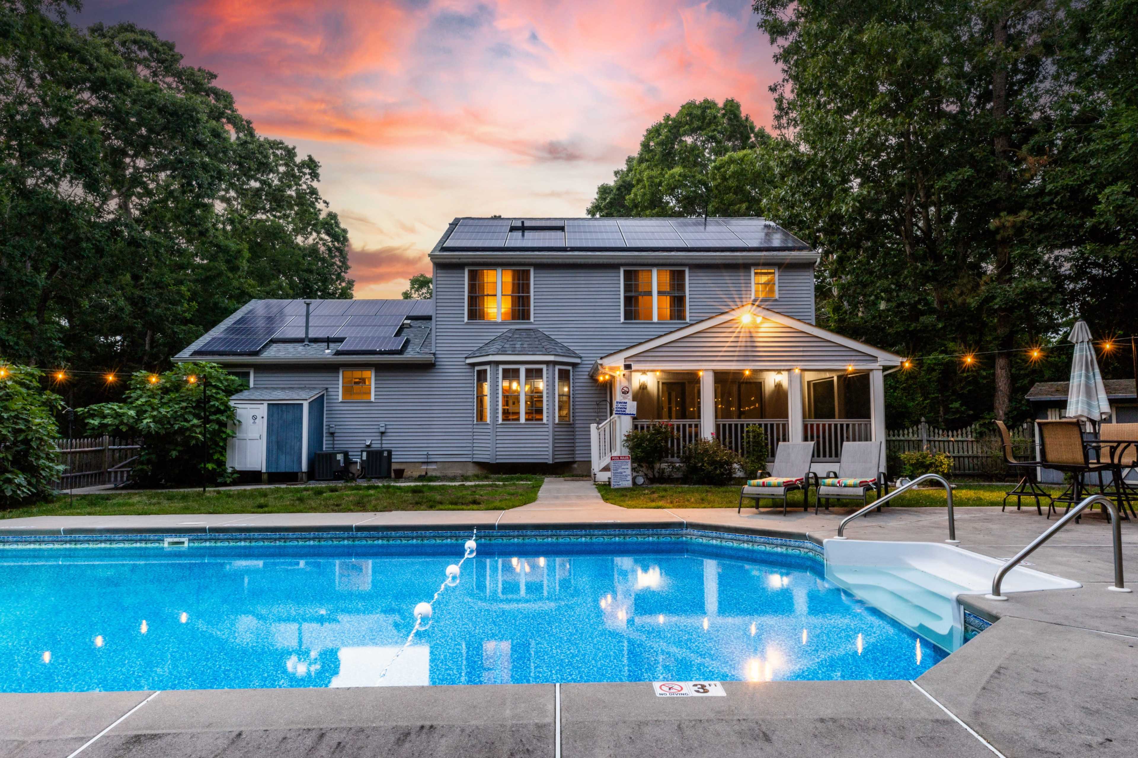 A two-story house with solar panels and a pool in the foreground, set against a colorful sunset sky.