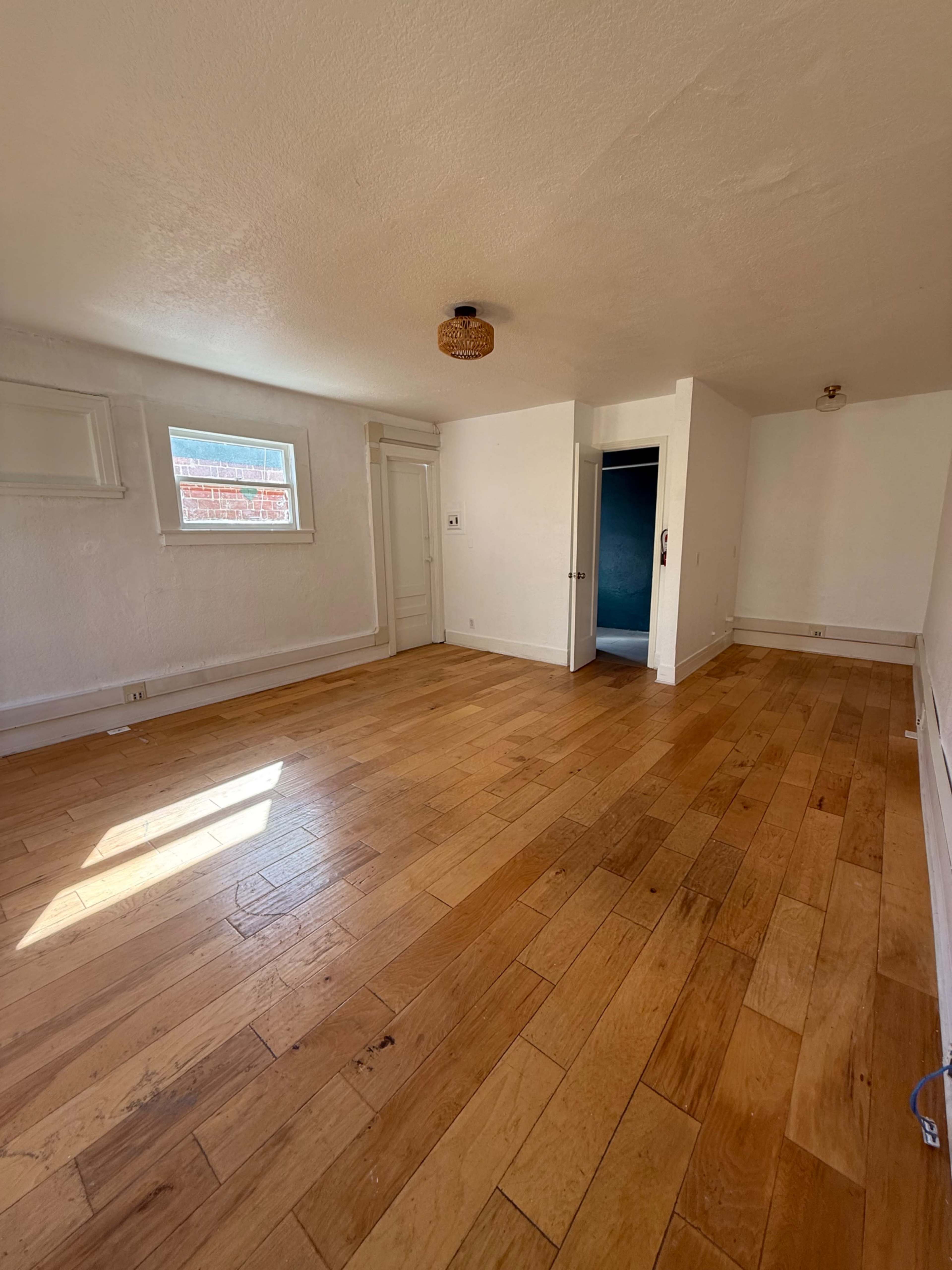 The image shows a vacant room with hardwood flooring, white walls, and a small window allowing natural light.