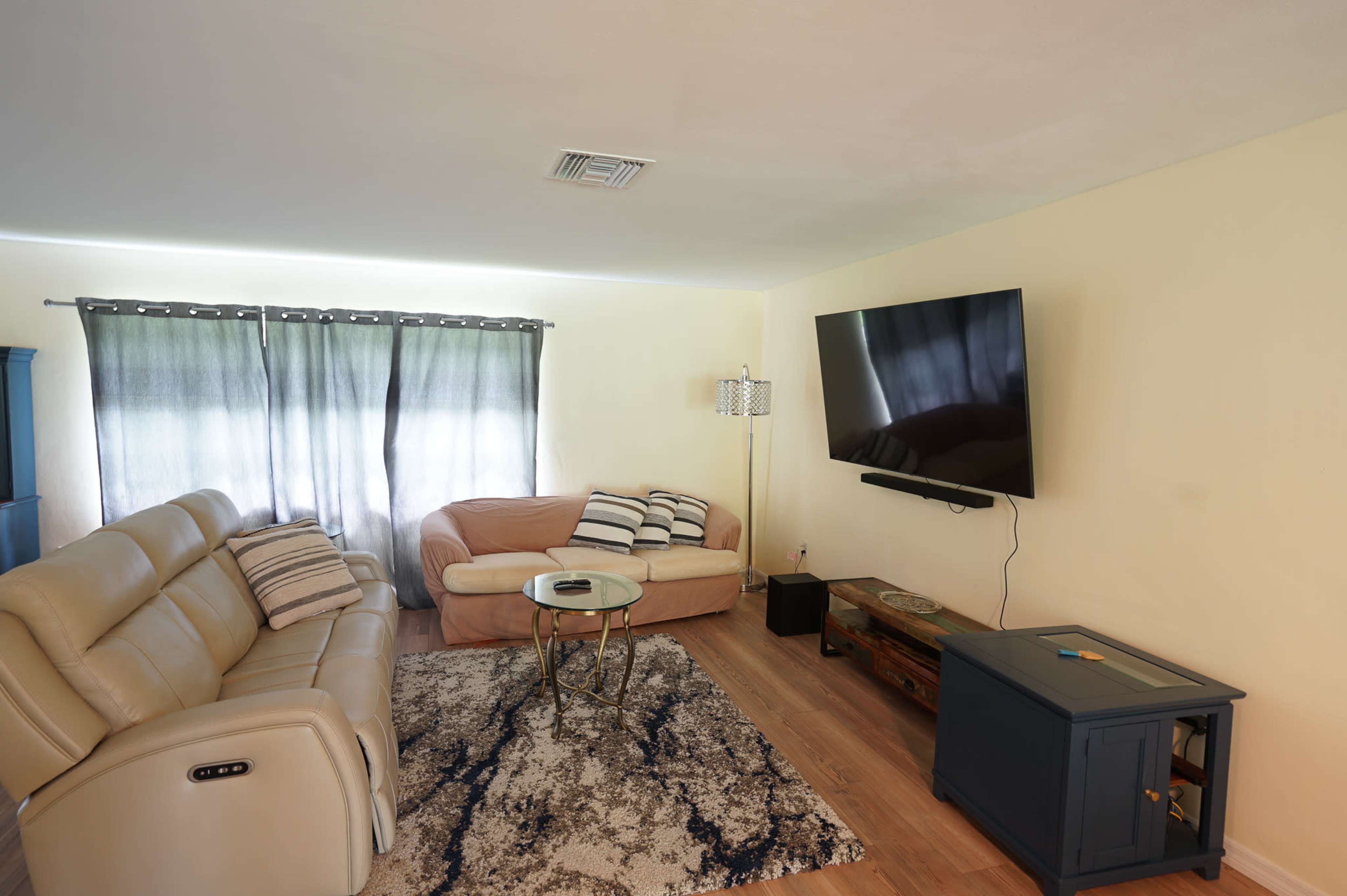A living room with a beige couch, a light-colored recliner, a glass coffee table, and a mounted television on the wall, accompanied by muted curtains and a wooden side table.