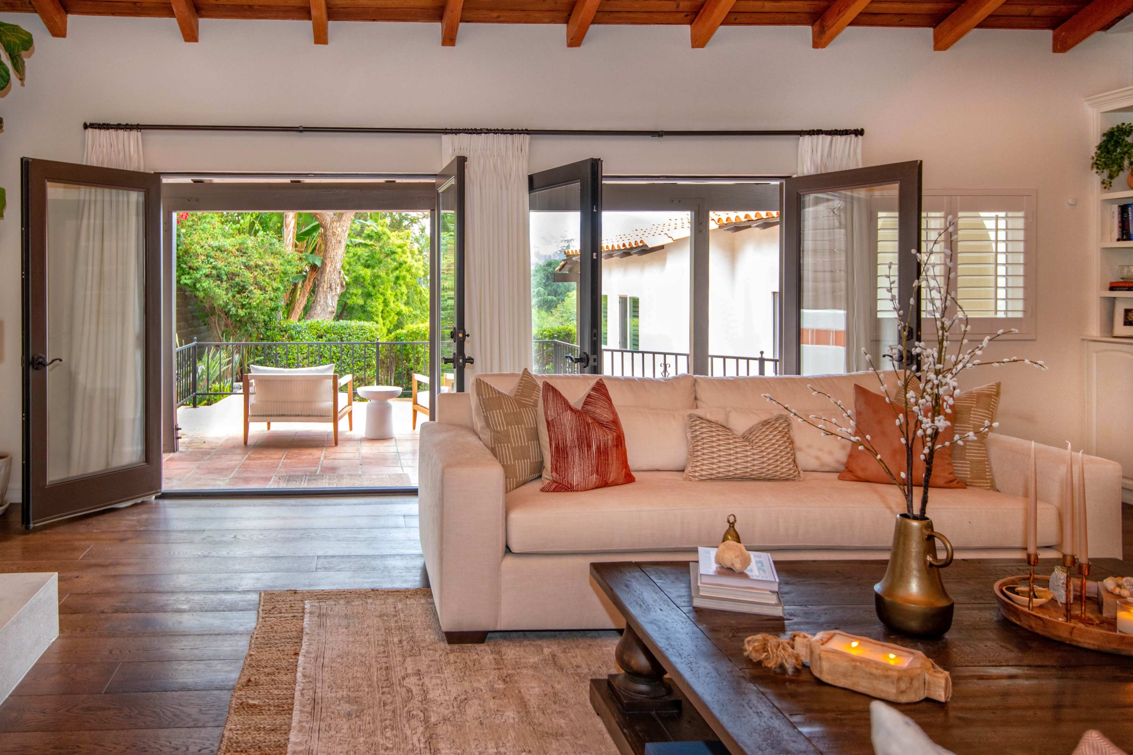 A well-lit living room features a beige sofa adorned with decorative pillows, facing a pair of open French doors that lead to a patio surrounded by greenery.