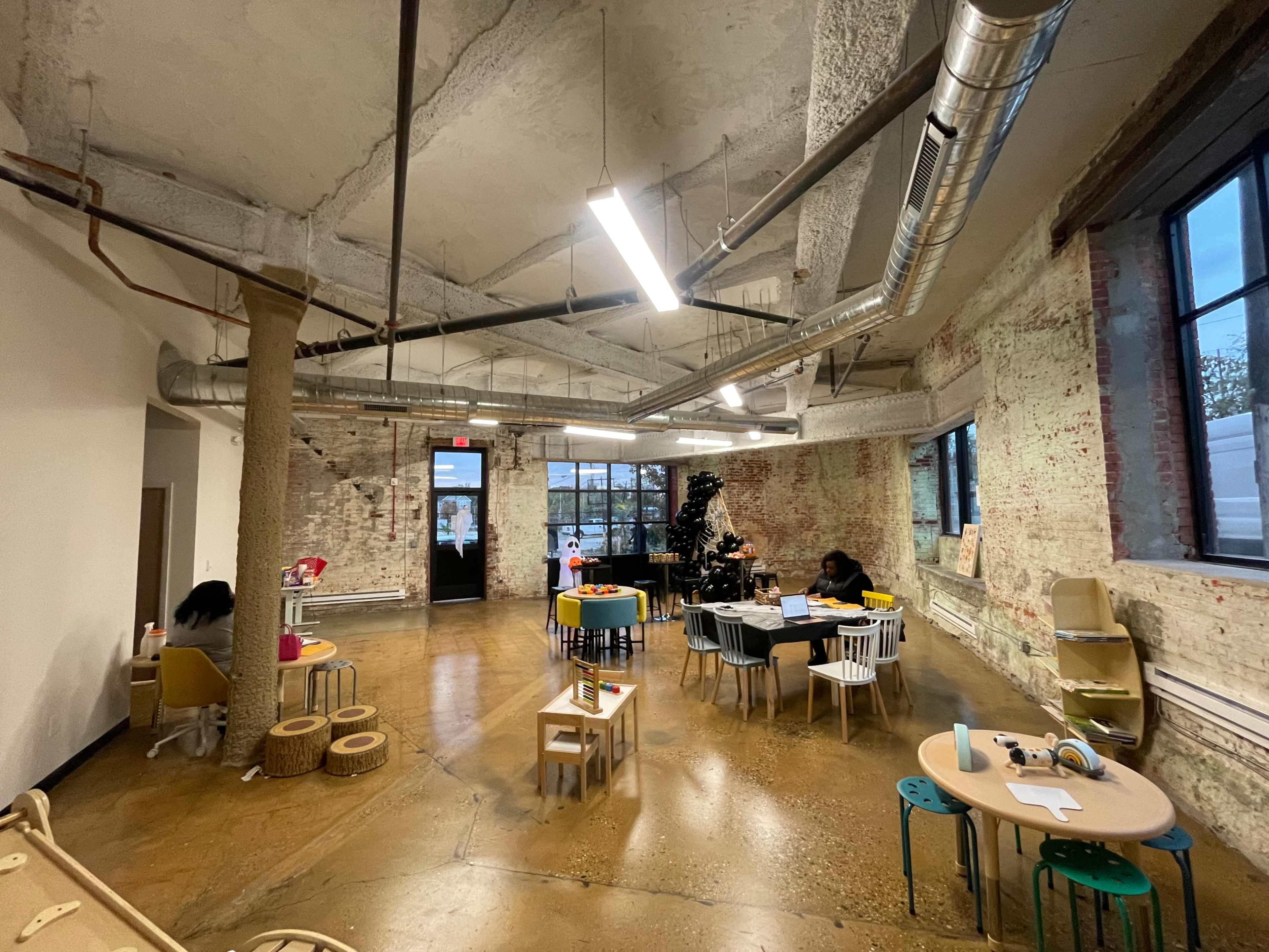 An open play area with various child-sized tables and chairs, exposed brick walls, and large windows providing natural light.
