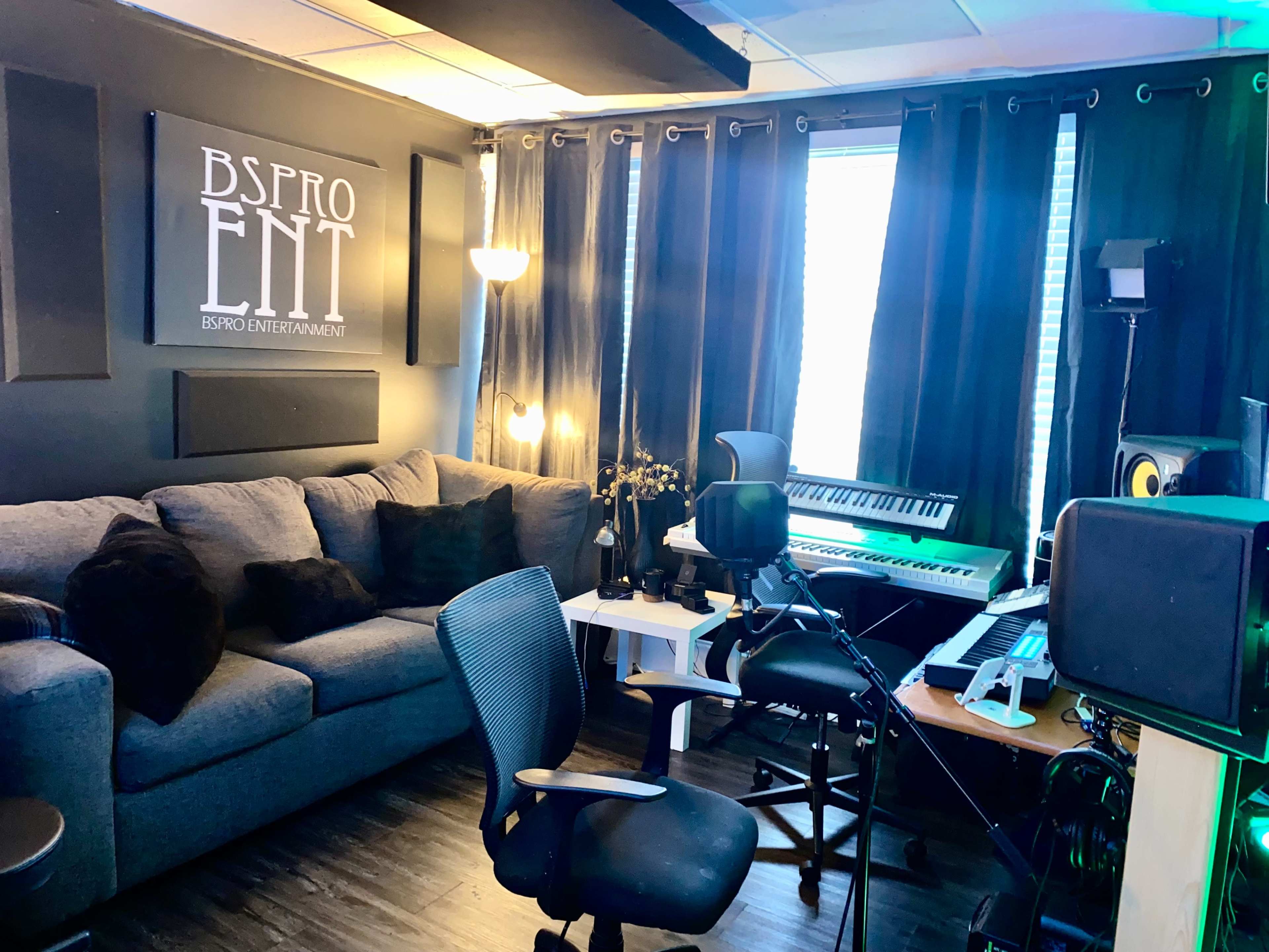 The image shows a music studio featuring a gray couch, a keyboard, audio equipment, and decorative curtains.