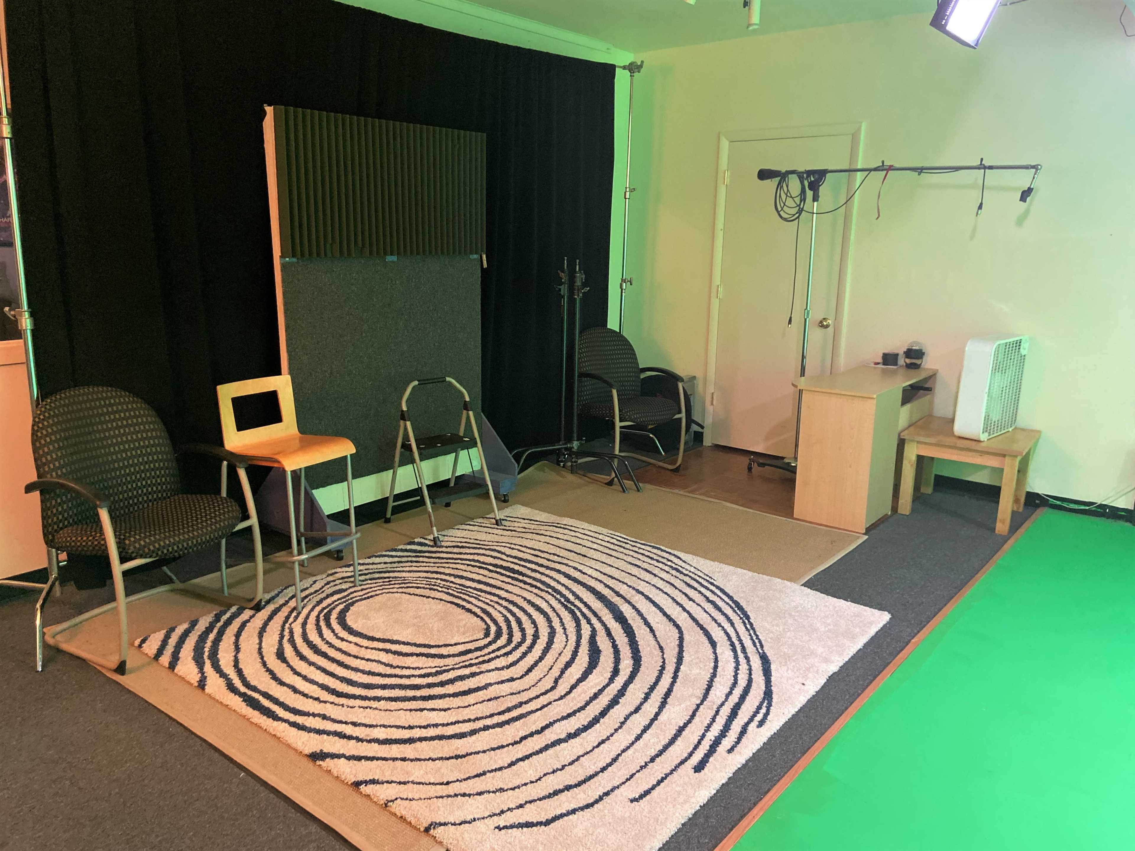 The image shows a small recording studio featuring a rug with concentric circles, two chairs, a wooden table, and a soundproof panel against a black curtain.