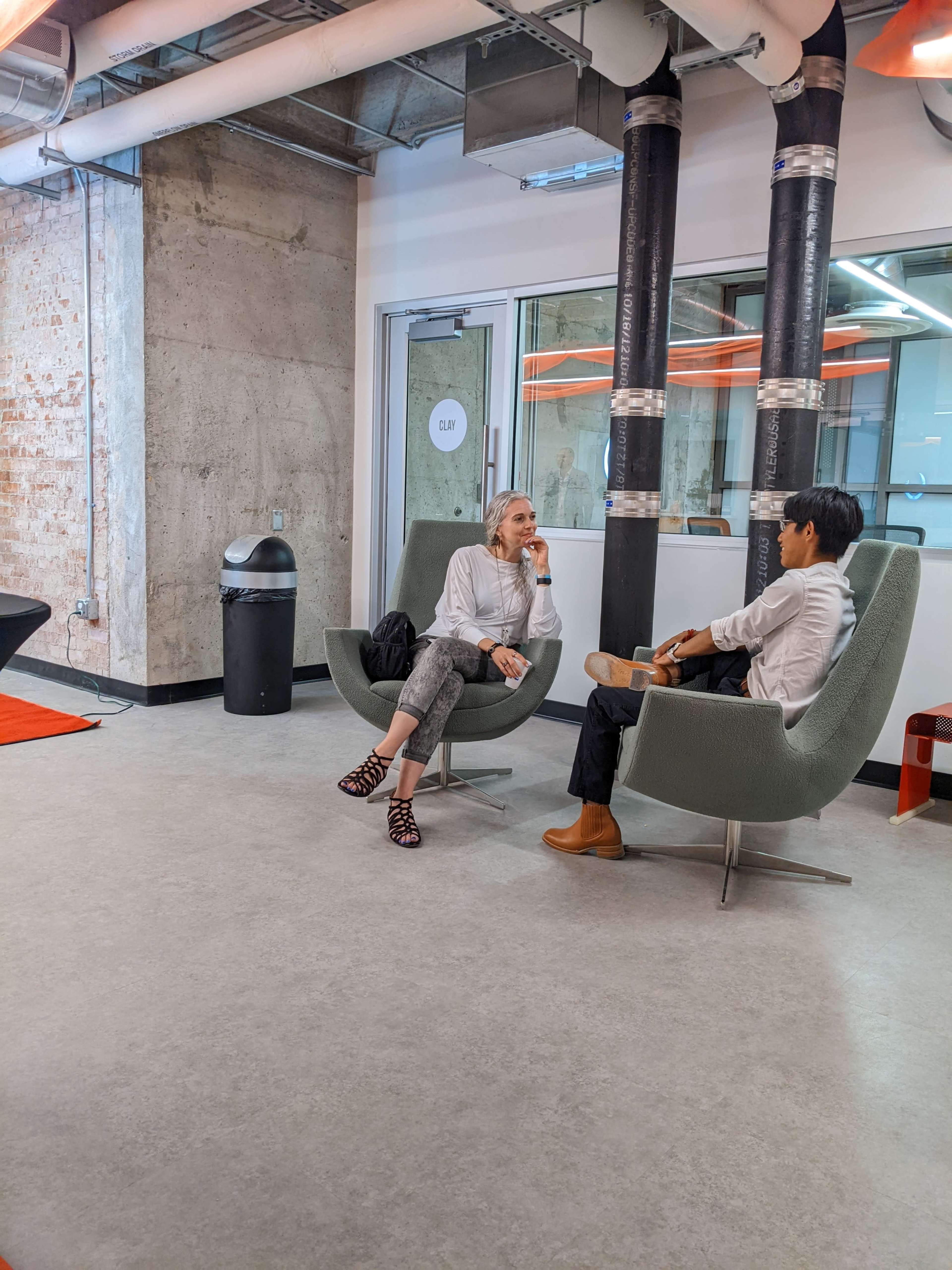 Two people sit in modern chairs in a spacious, industrial-style room with exposed concrete walls and large windows.