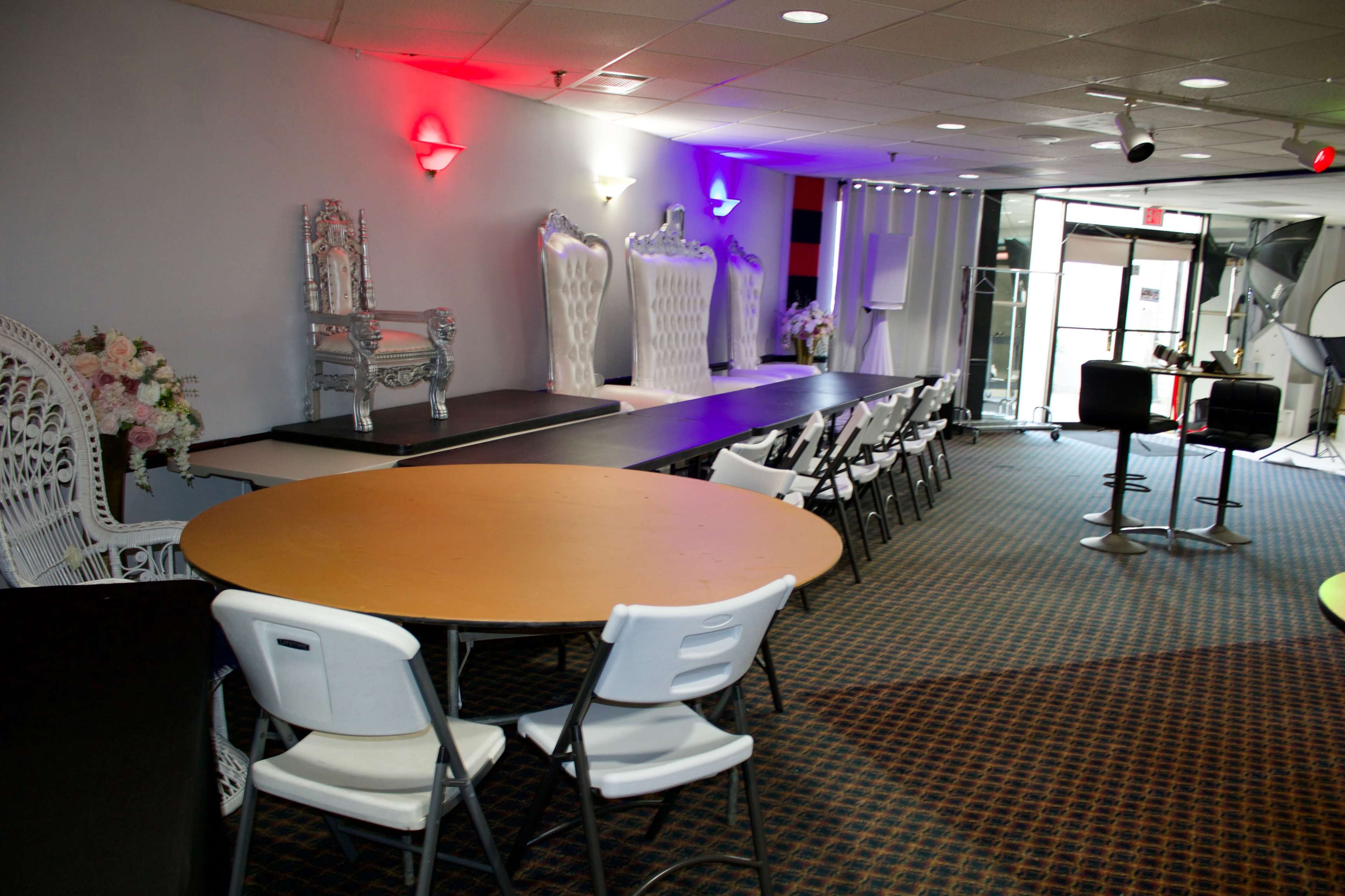 The image shows a spacious event room with a long table and several white chairs arranged around it, featuring decorative elements like a silver throne and colorful lighting.