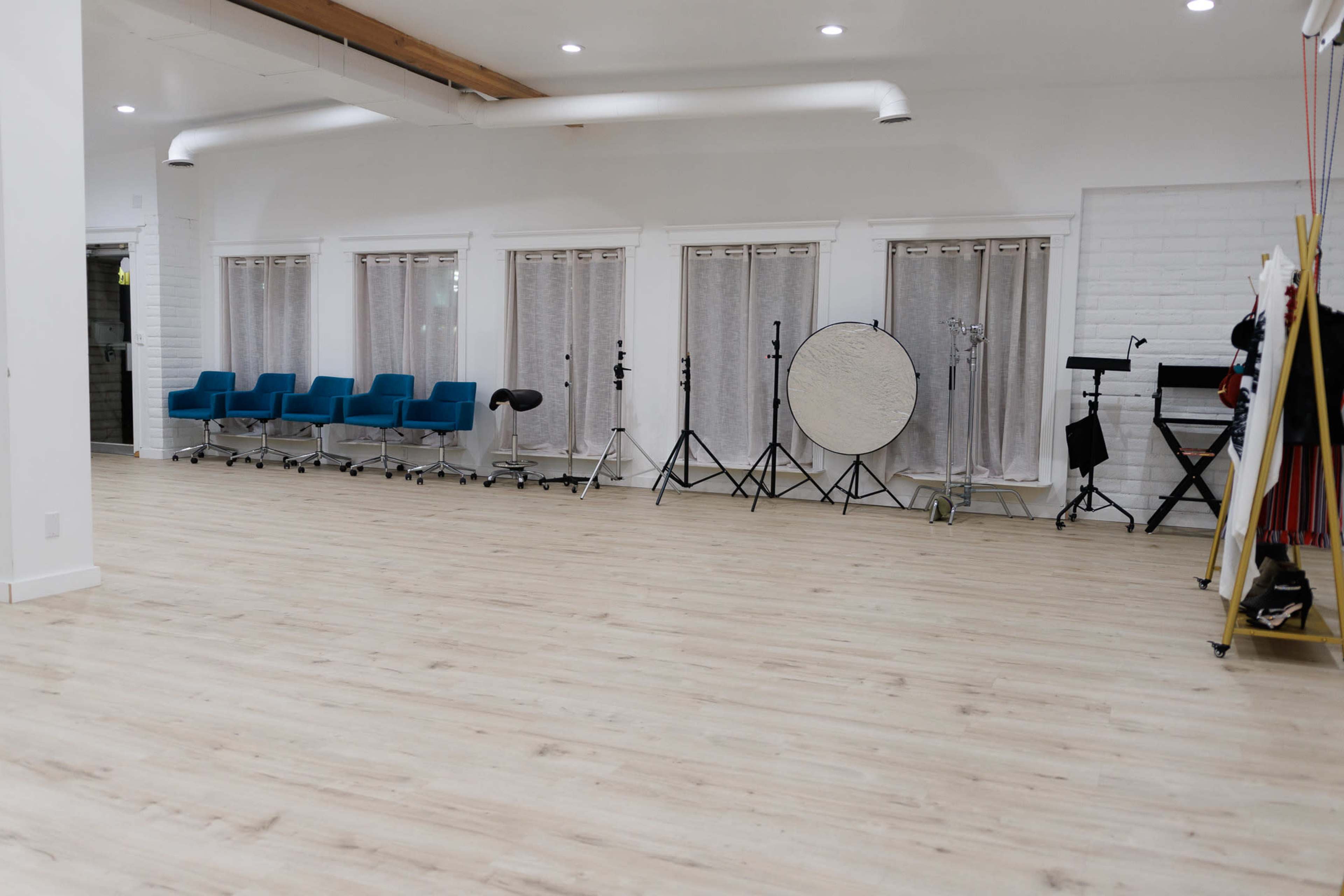 The image shows a spacious studio with a light wooden floor, featuring blue chairs, musical equipment, and several windows covered with curtains.