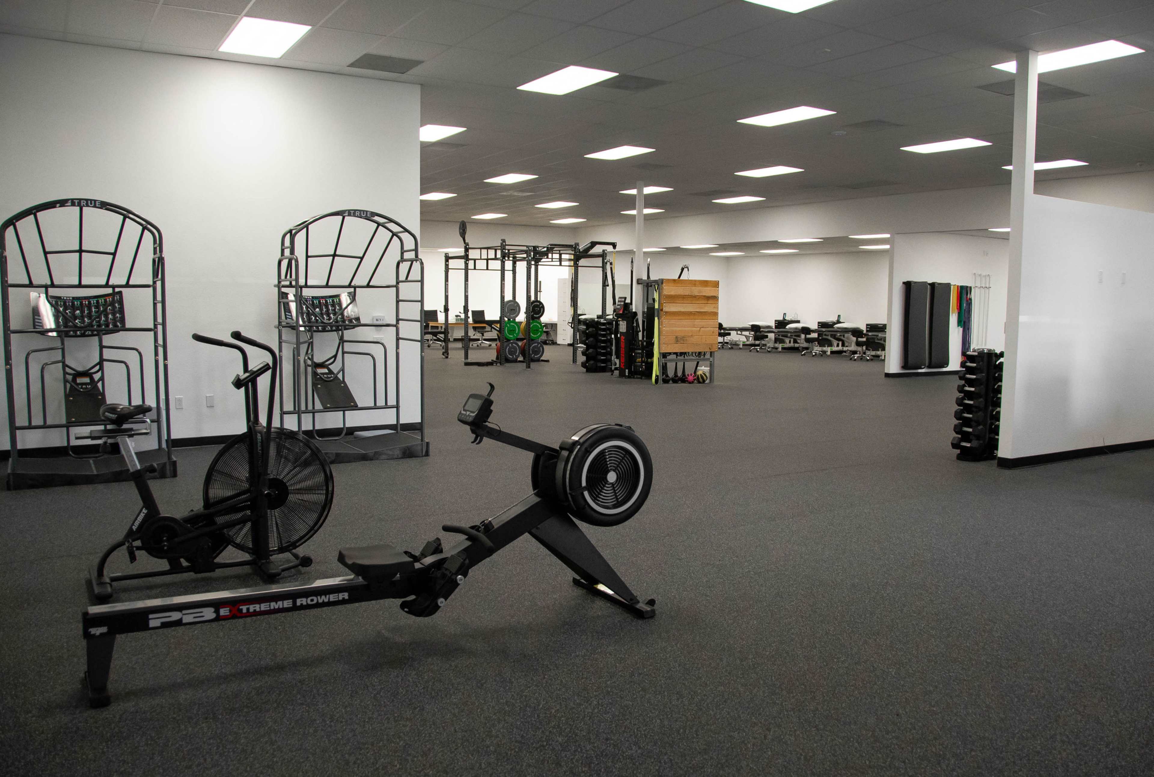 A spacious gym interior with exercise equipment, including a rowing machine and a bike, set against a backdrop of weightlifting stations and open training areas.