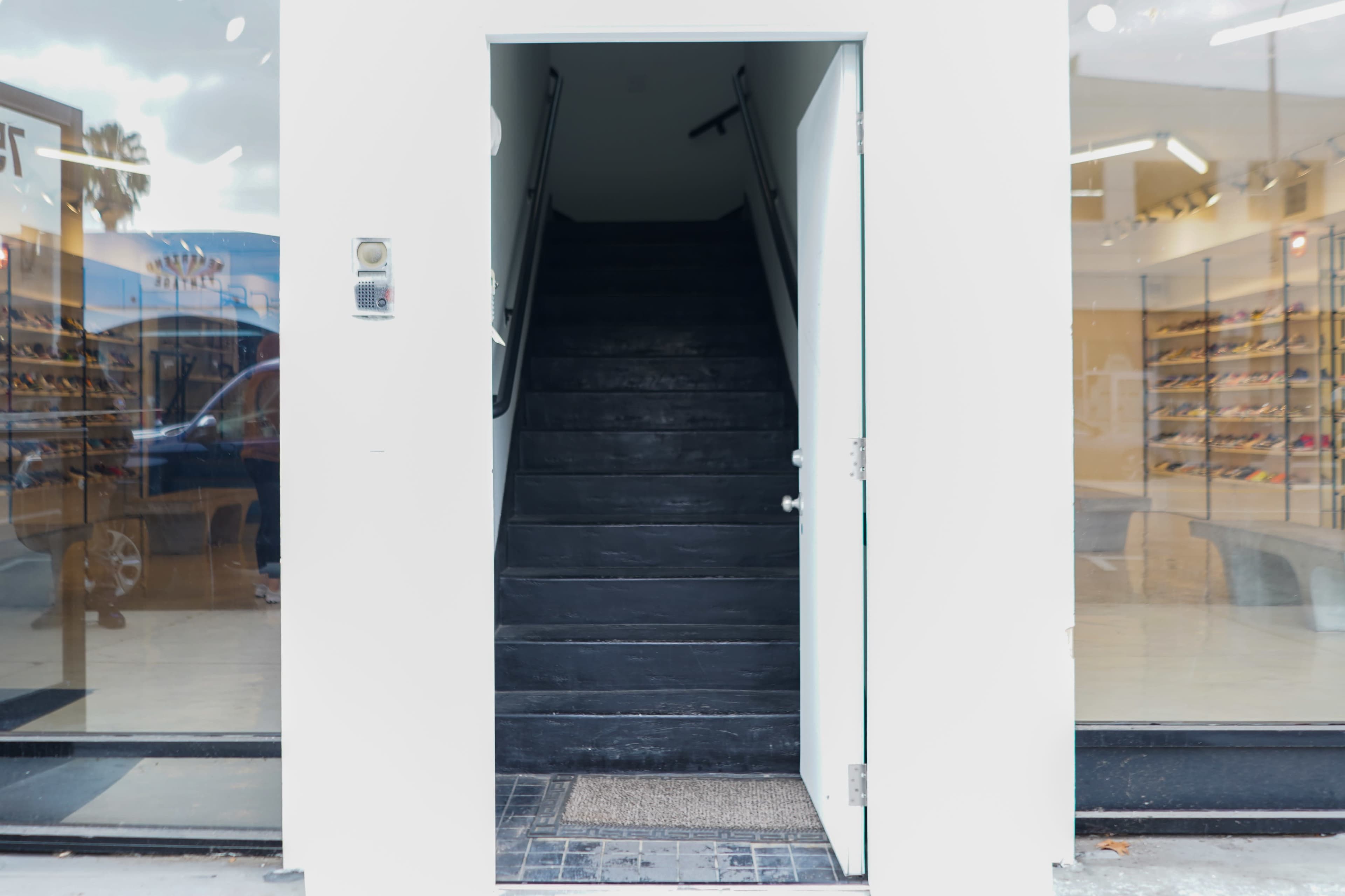 A set of black stairs leads up from an open door in a white wall, flanked by large glass windows.
