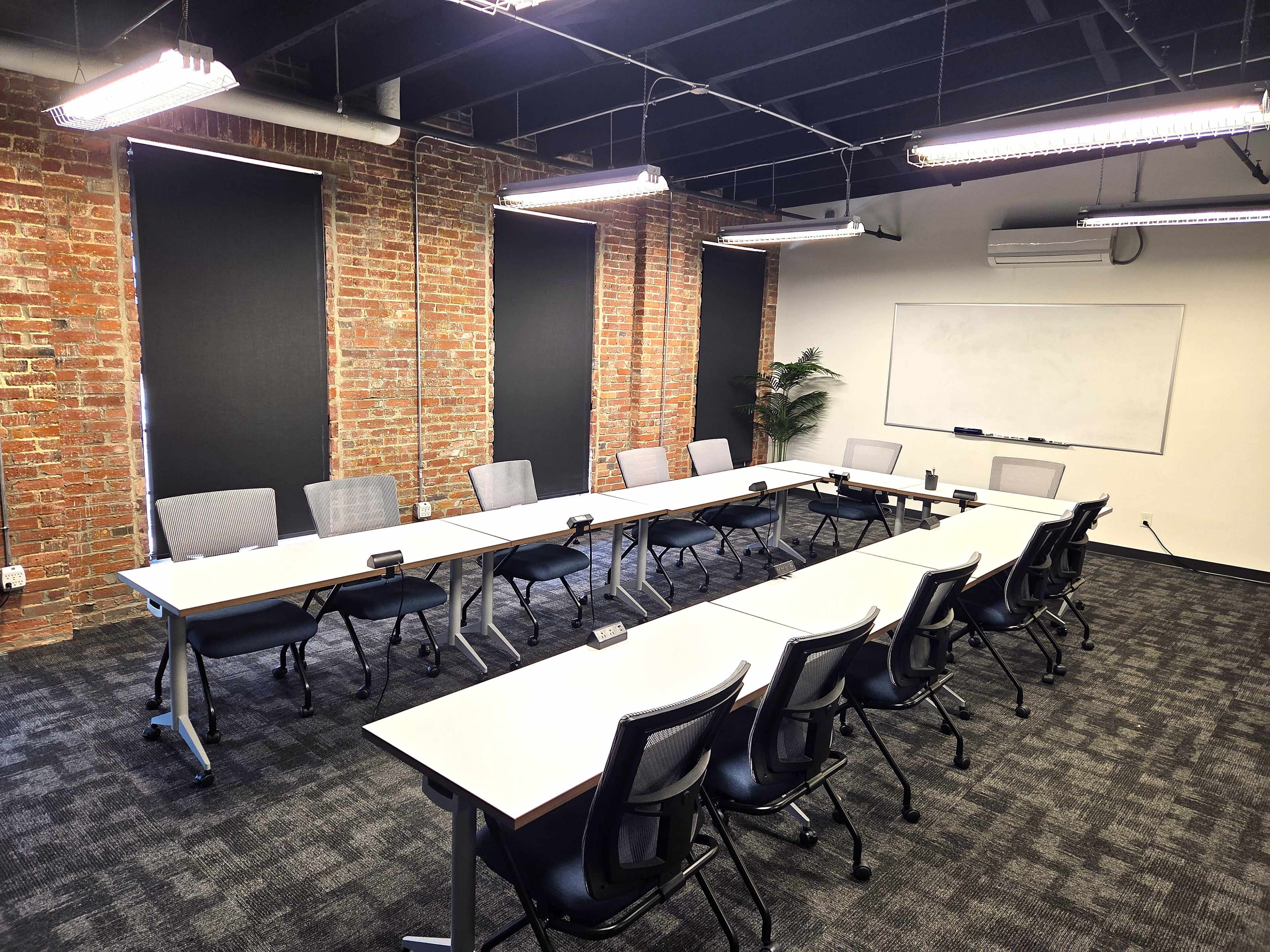 The image depicts a modern conference room with a long table surrounded by chairs, exposed brick walls, and a whiteboard at one end.