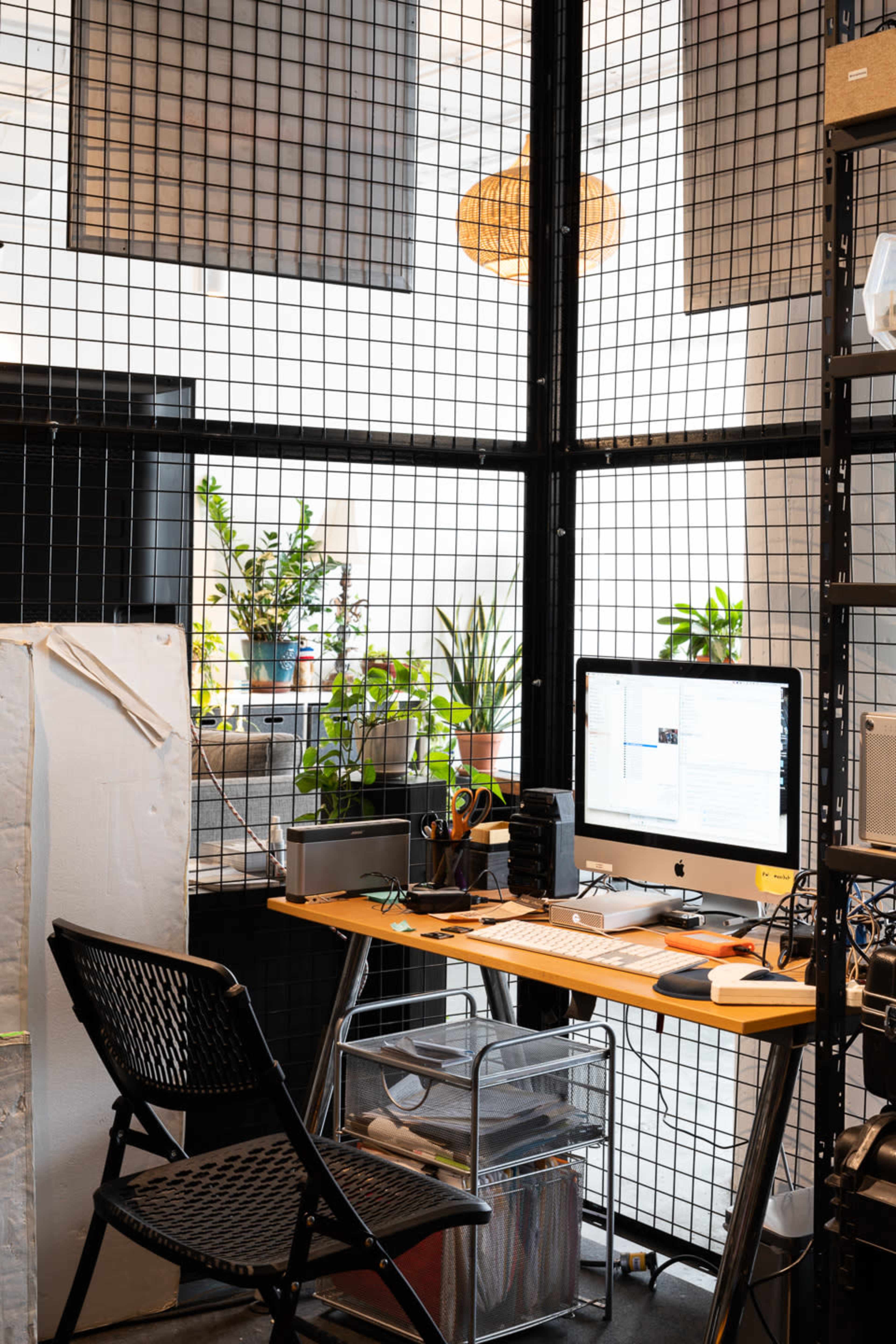 A modern office workspace features a wooden desk with a computer, surrounded by plants and enclosed by a grid-like metal structure.