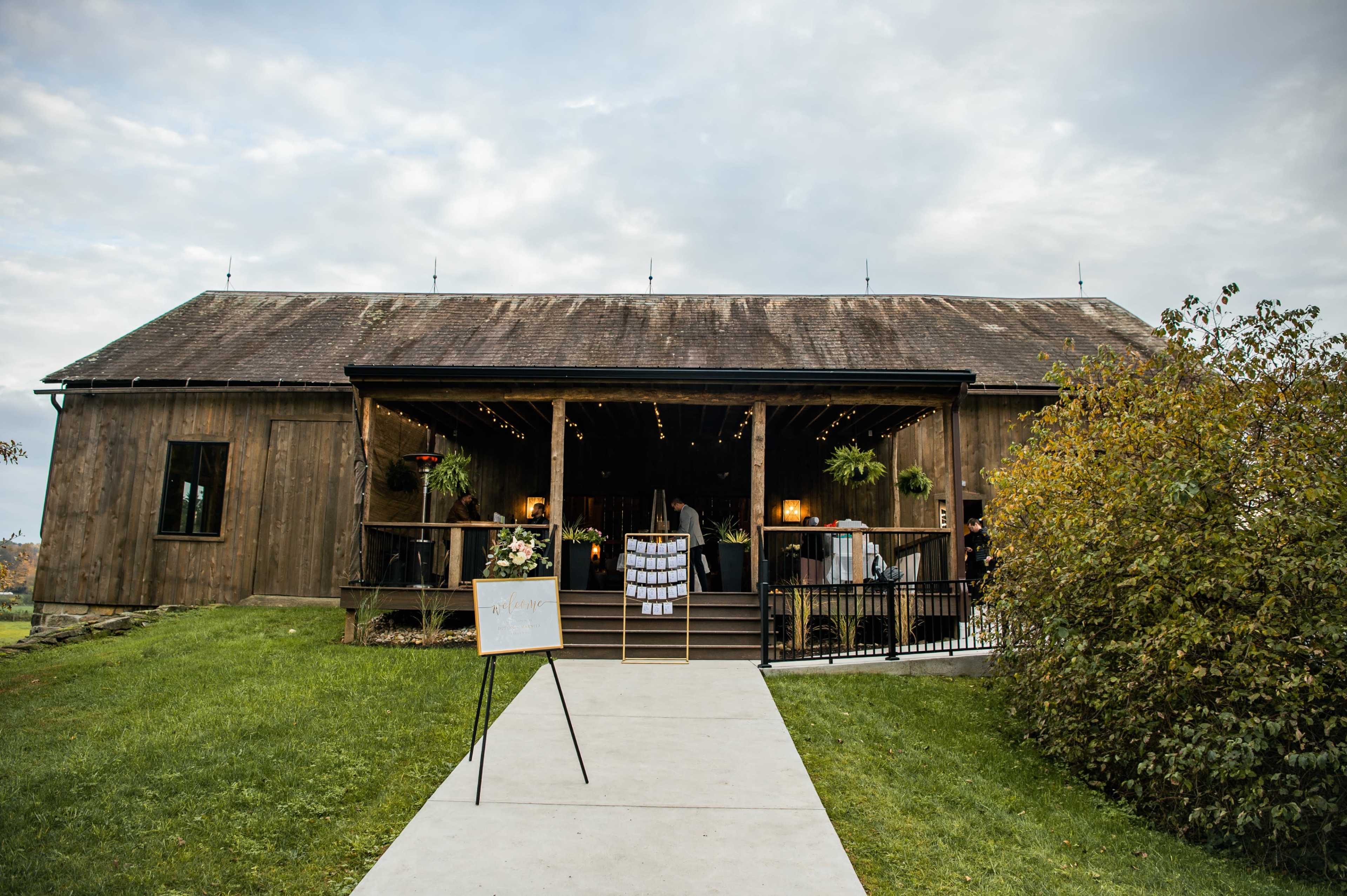 A wooden barn with a large entrance and steps is surrounded by greenery and has a sign in front.