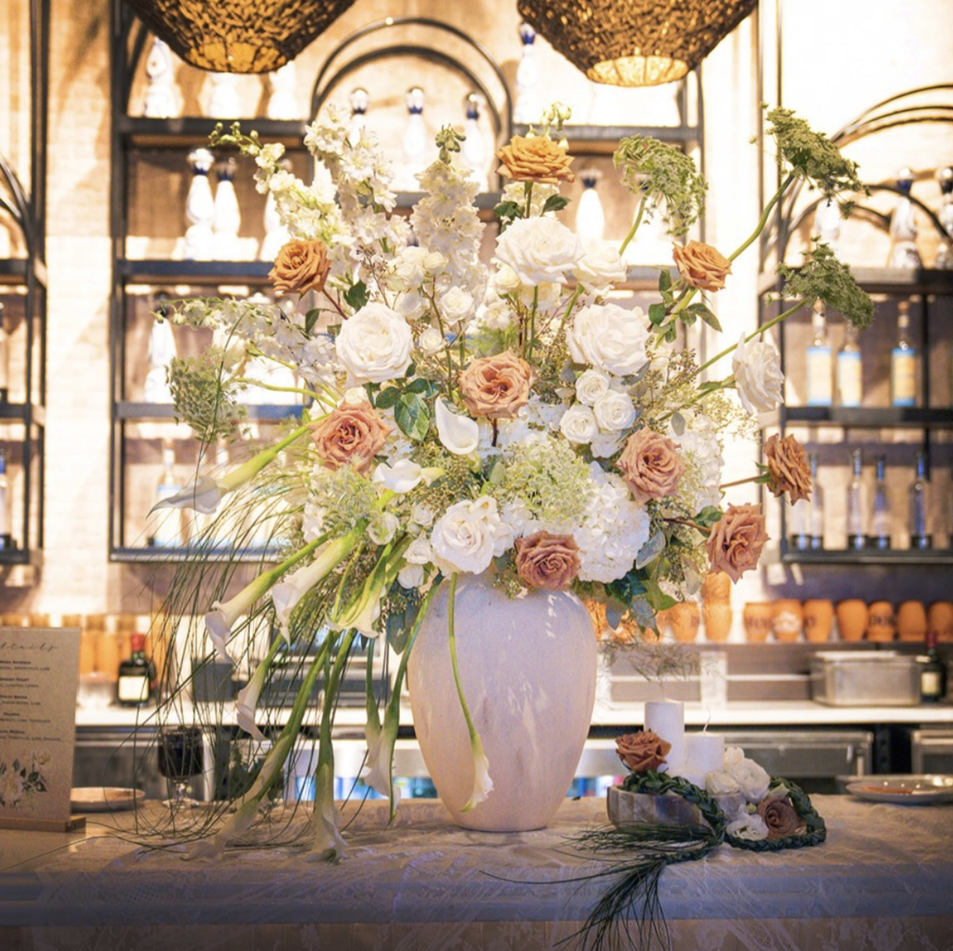 A large, elegant floral arrangement featuring white and peach roses, greenery, and other assorted flowers is displayed in a vase on a table in a restaurant setting.
