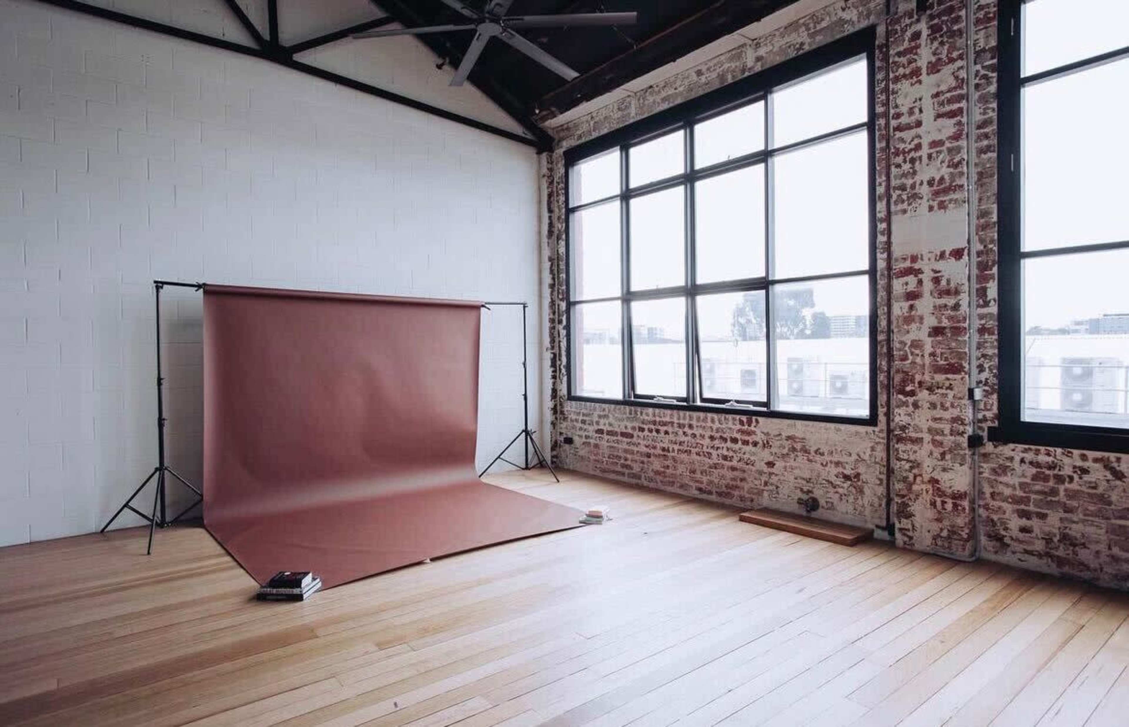 An empty studio space with large windows, a brick wall, wooden flooring, and a brown backdrop setup for photography.