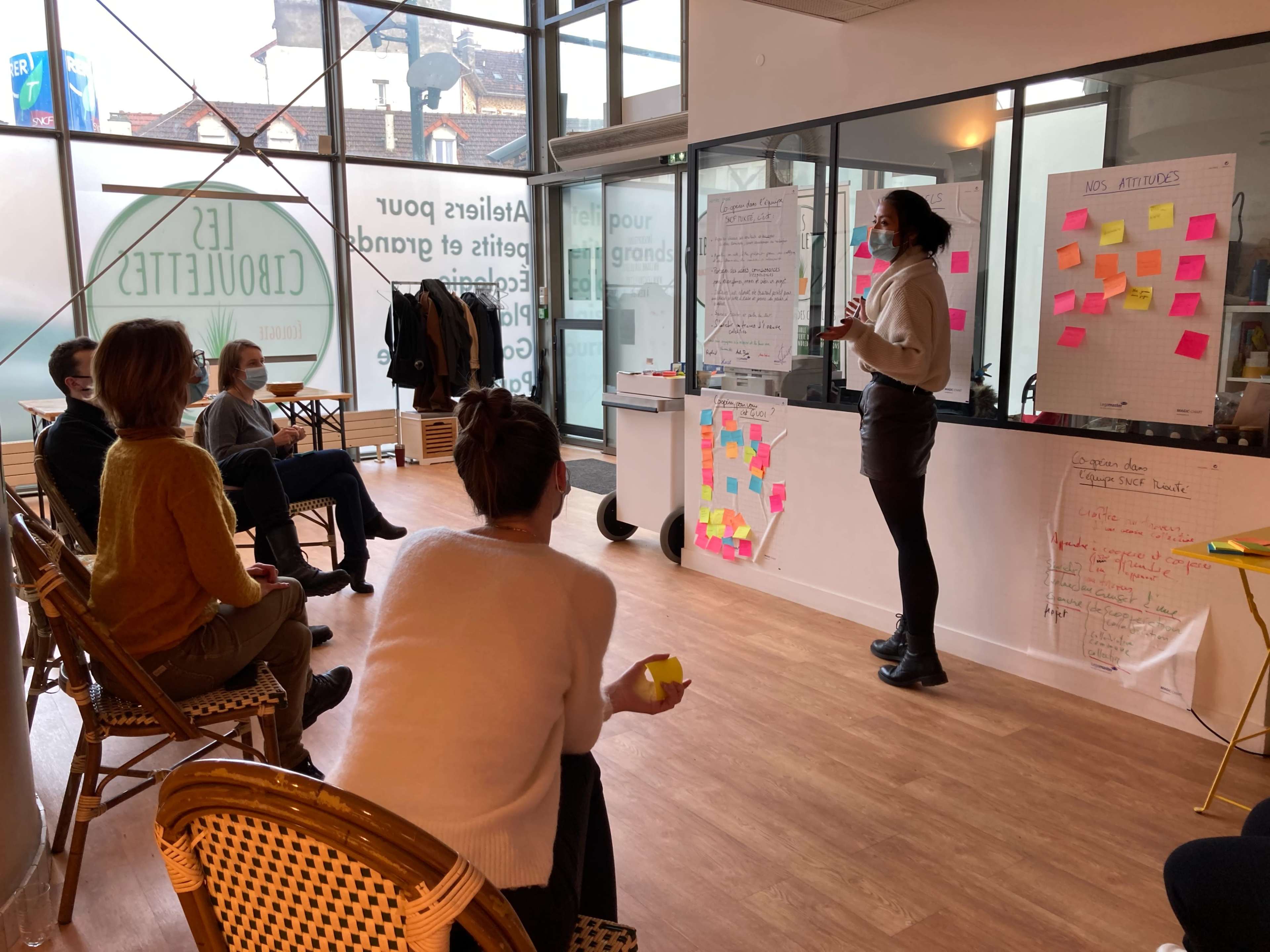 A group of people is seated in a brightly lit room, attentively listening to a presenter who stands by a board filled with colorful sticky notes.