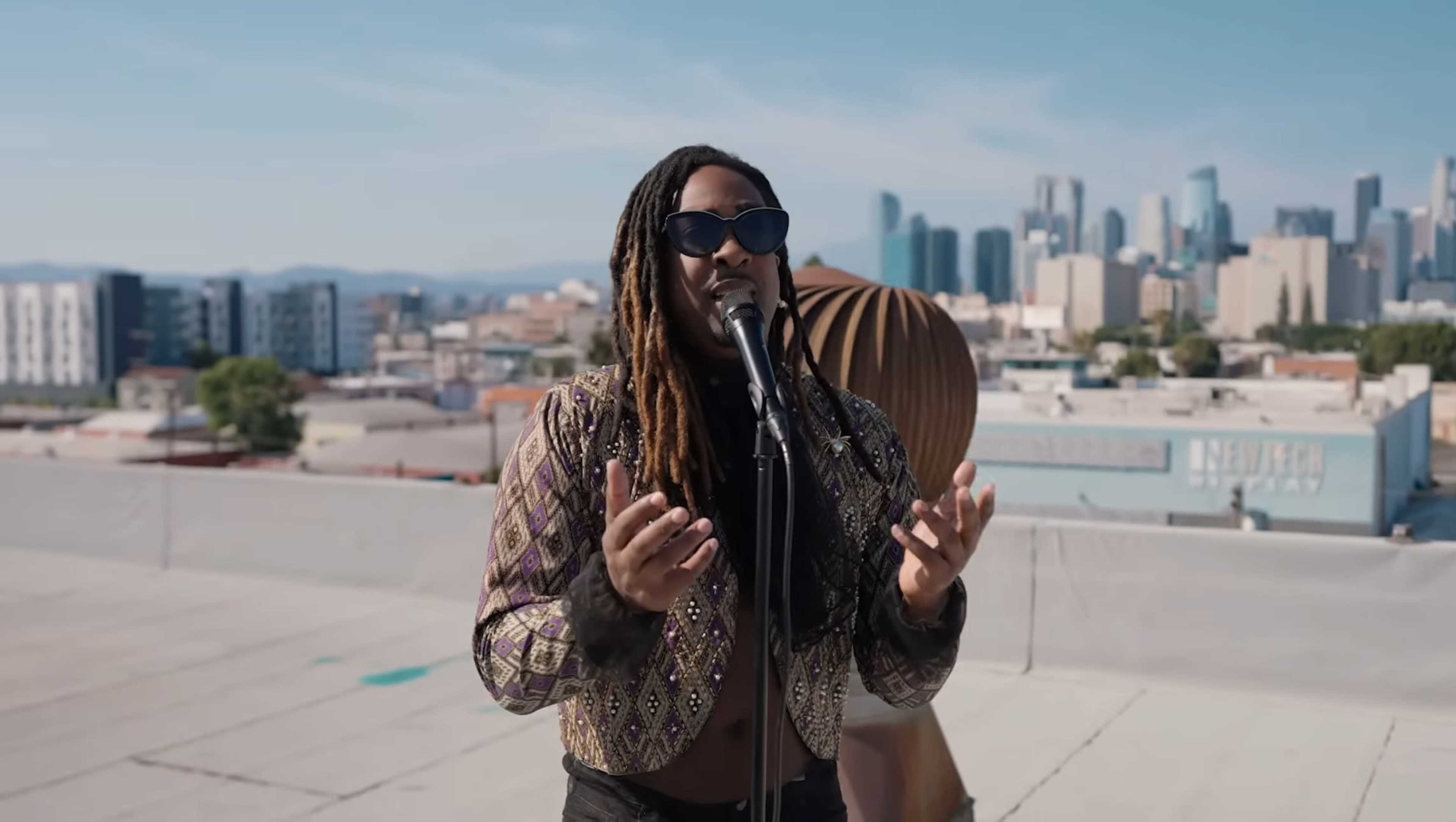 A musician with long, braided hair performs on a rooftop, microphone in hand, with a city skyline in the background.