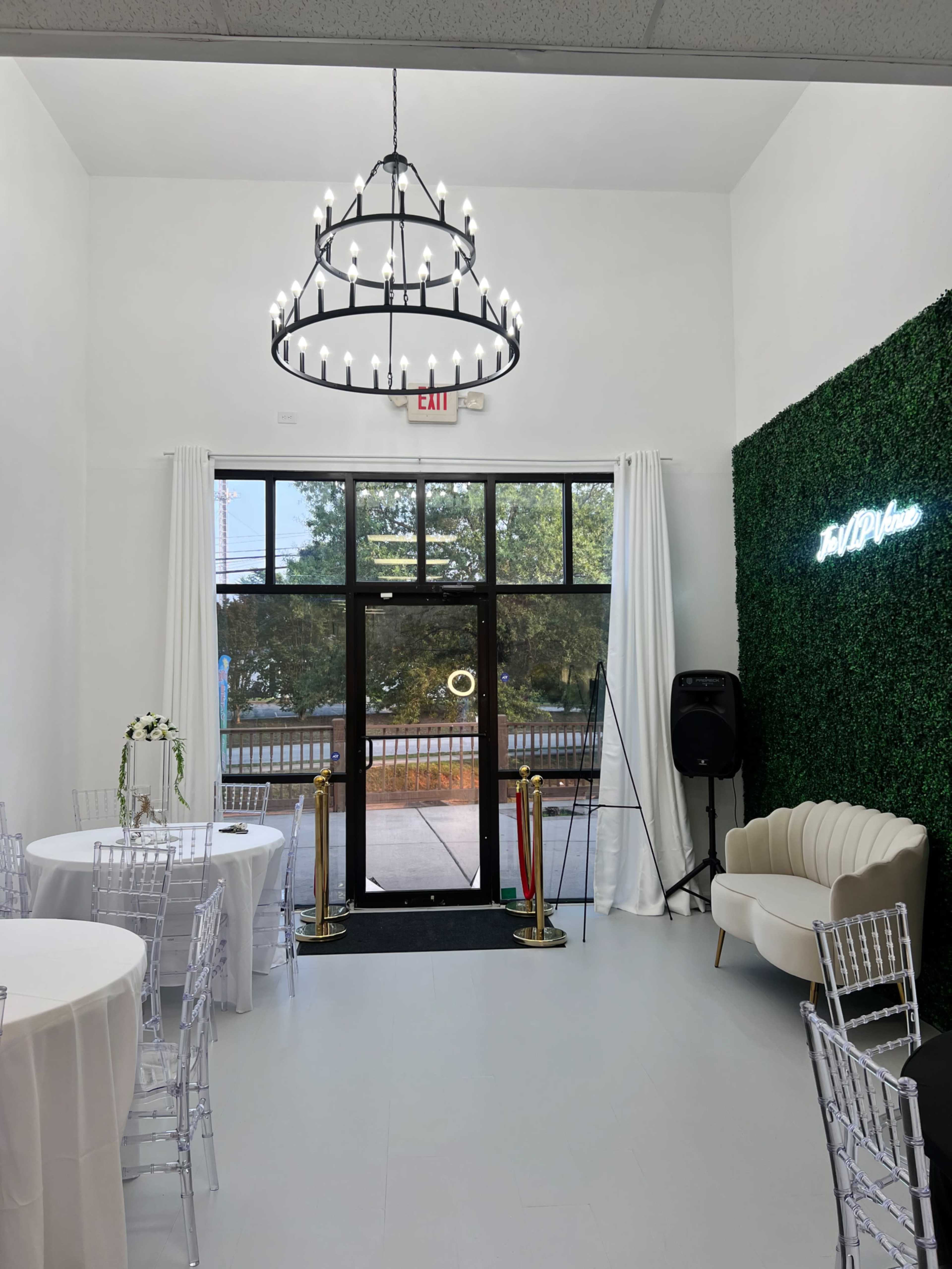 The image shows a well-lit event space featuring a large chandelier, round tables with white tablecloths, and a green wall with the text "Full Bloom."