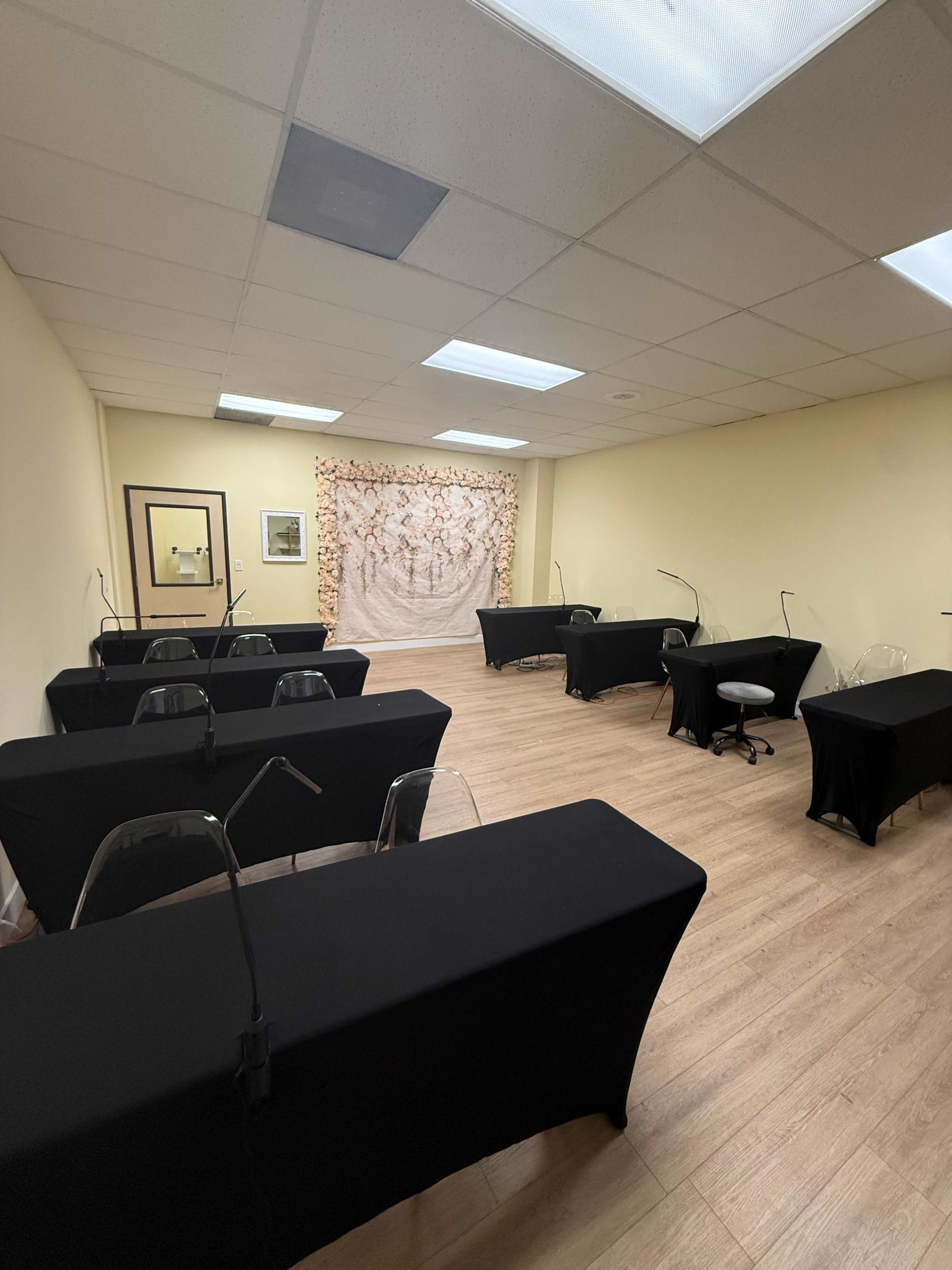 The image shows a spacious room arranged with several black-covered tables facing a decorative backdrop, with clear chairs and a single rolling stool.