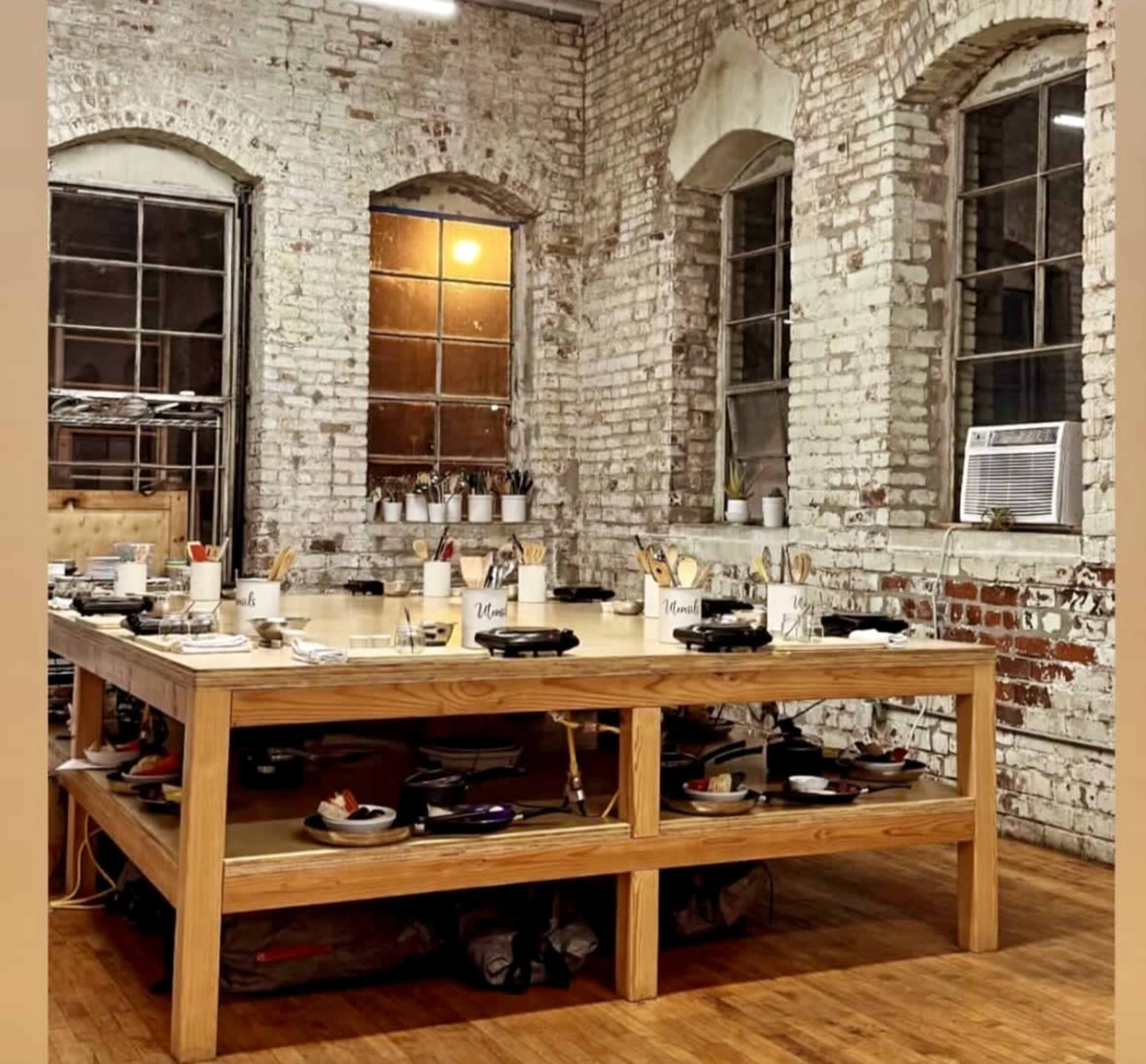 The image shows a large wooden table in a rustic room with exposed brick walls and multiple windows.