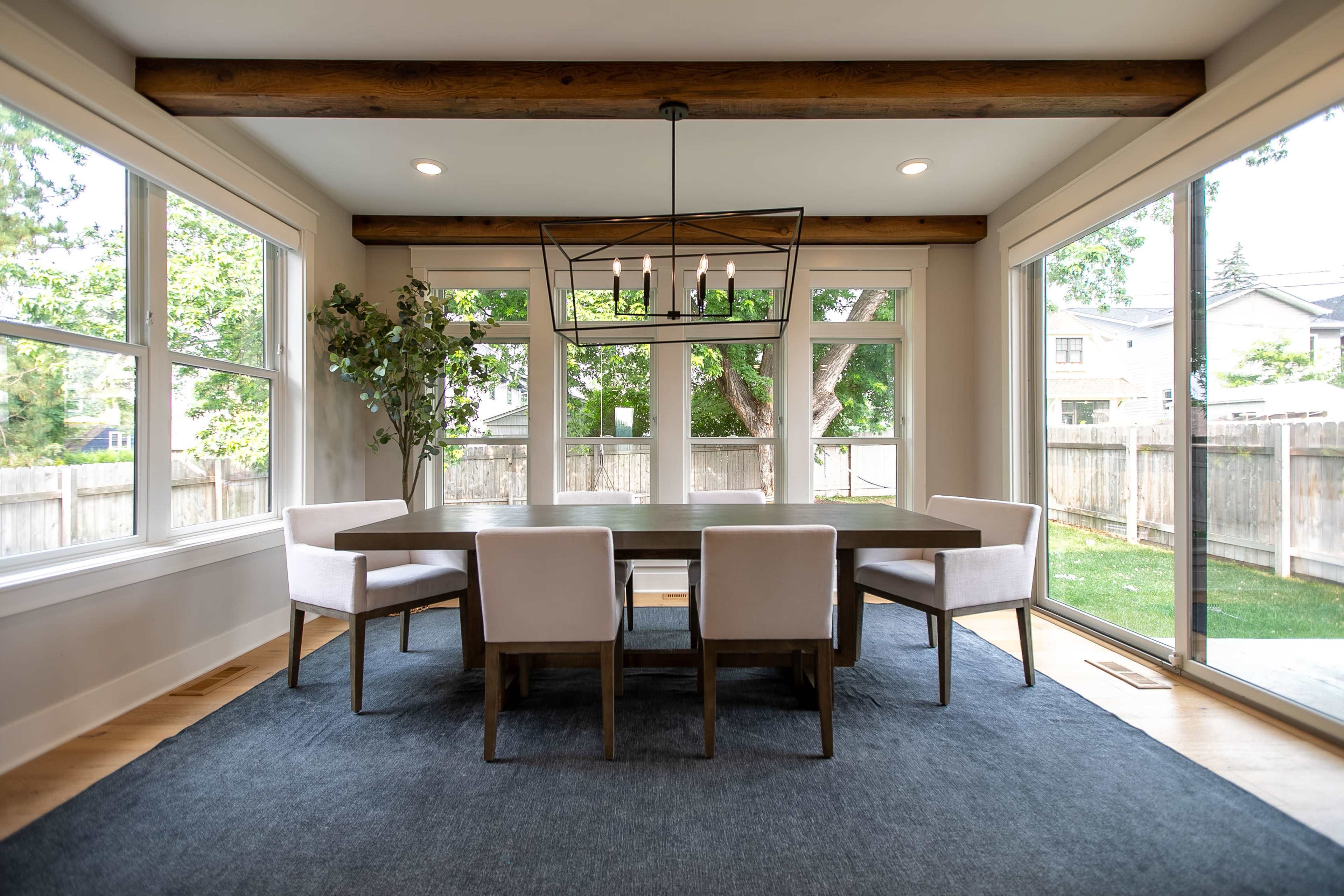 A modern dining room features a large wooden table surrounded by upholstered chairs, with large windows allowing natural light to illuminate the space.
