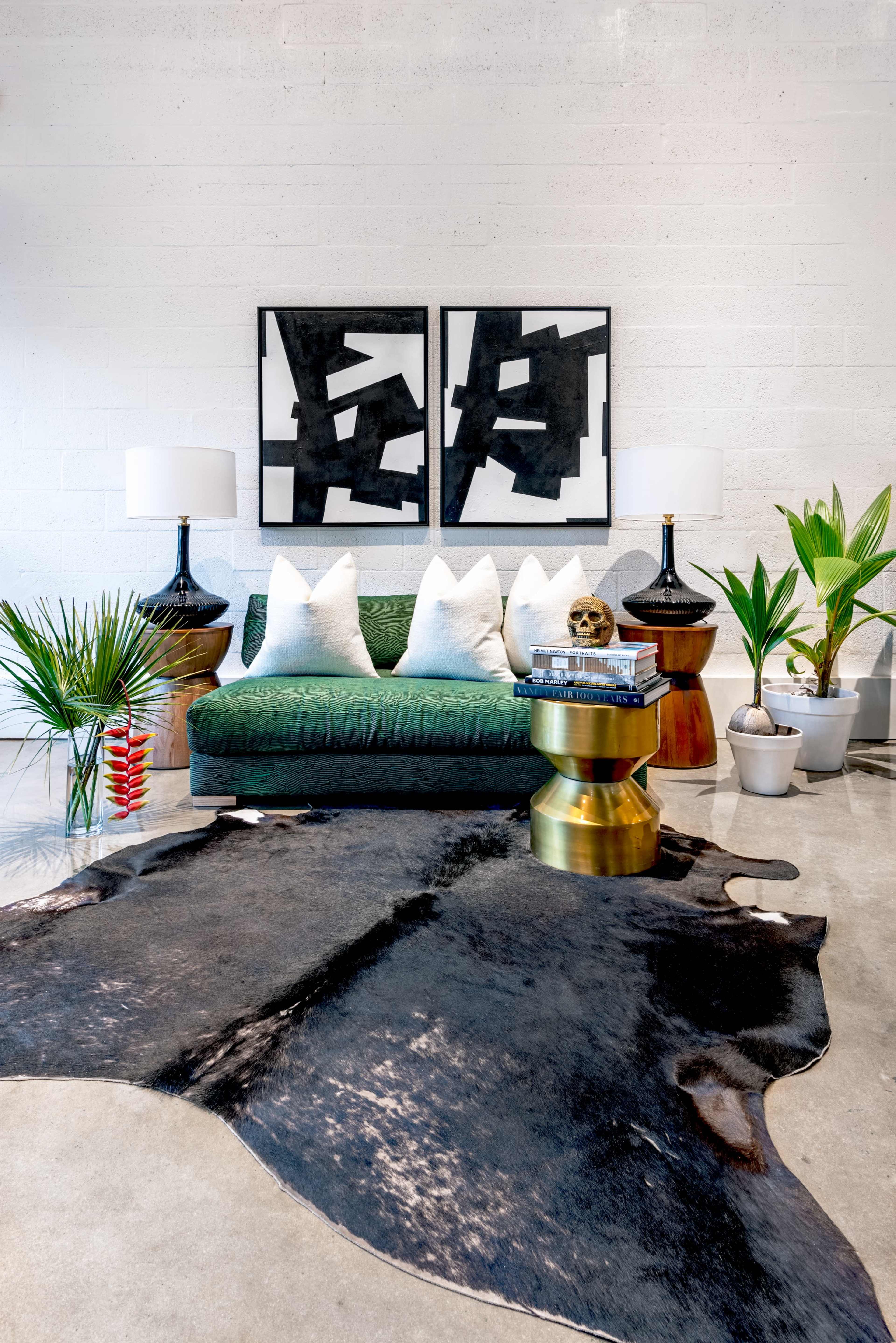 A modern living room with a green sofa adorned with white pillows, two black lamps on side tables, and a patterned area rug on a polished floor, surrounded by indoor plants and artwork on the walls.