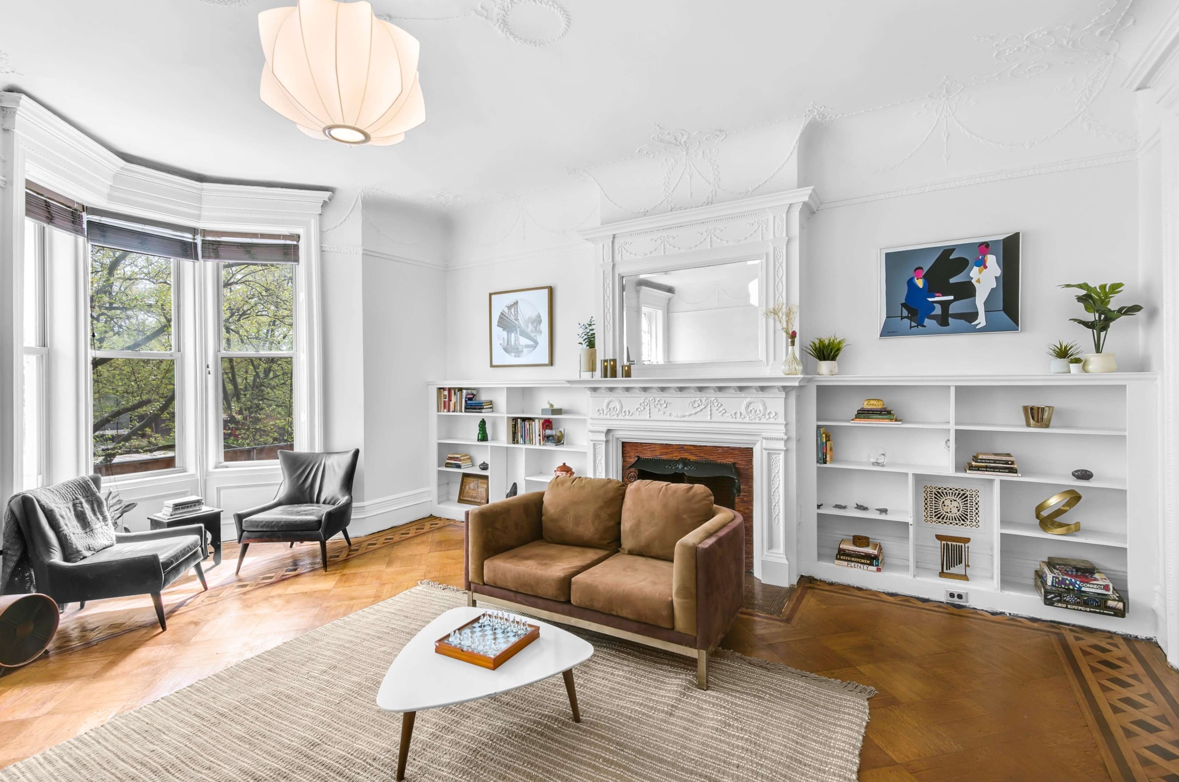 A light-filled living room features a brown sofa, a small white coffee table with a chess set, and a bookshelf against a wall, with a window showing greenery outside.