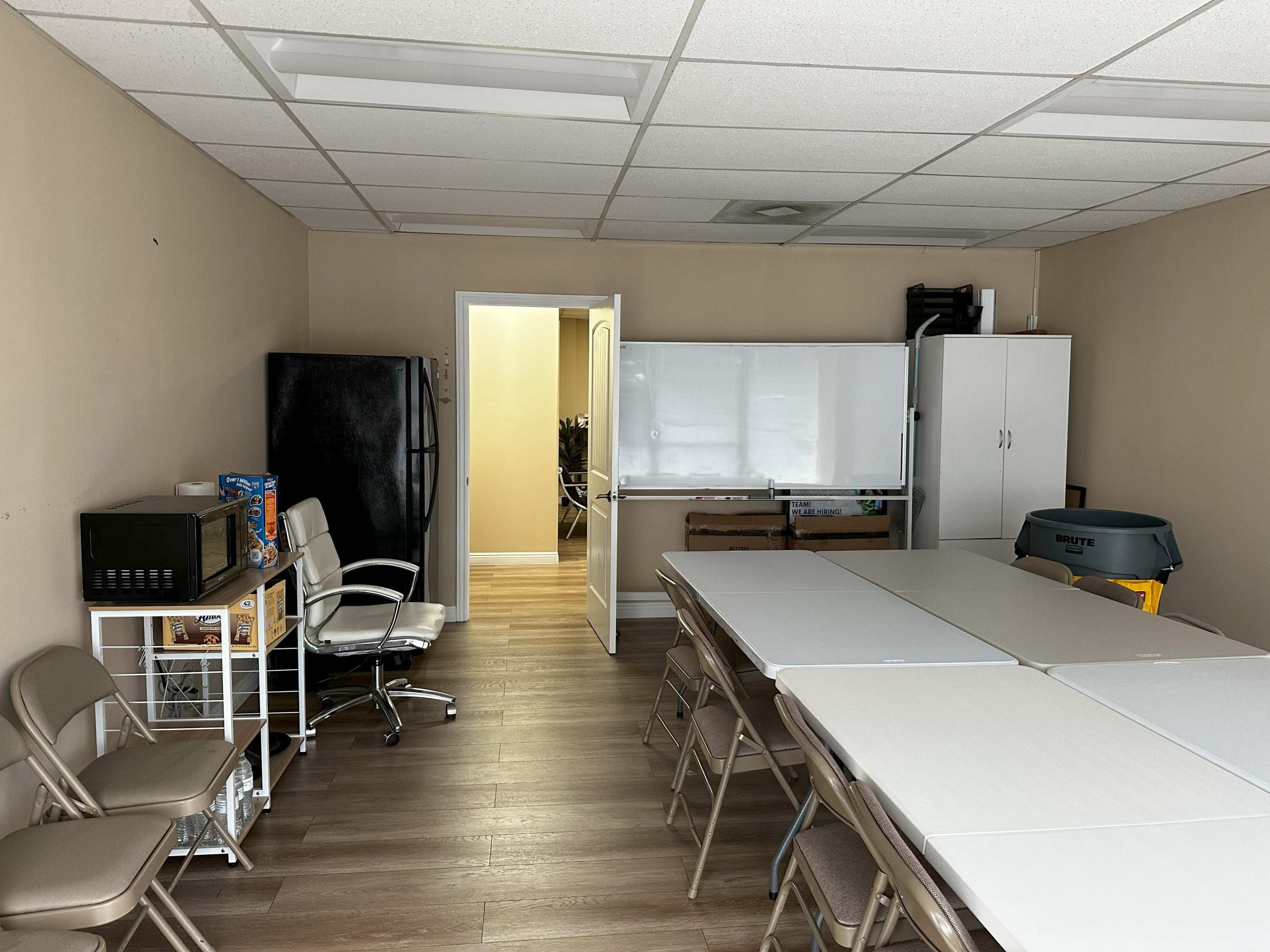 The image shows a neatly organized room featuring tables and chairs set up for a meeting, with a kitchenette area and a whiteboard on one wall.