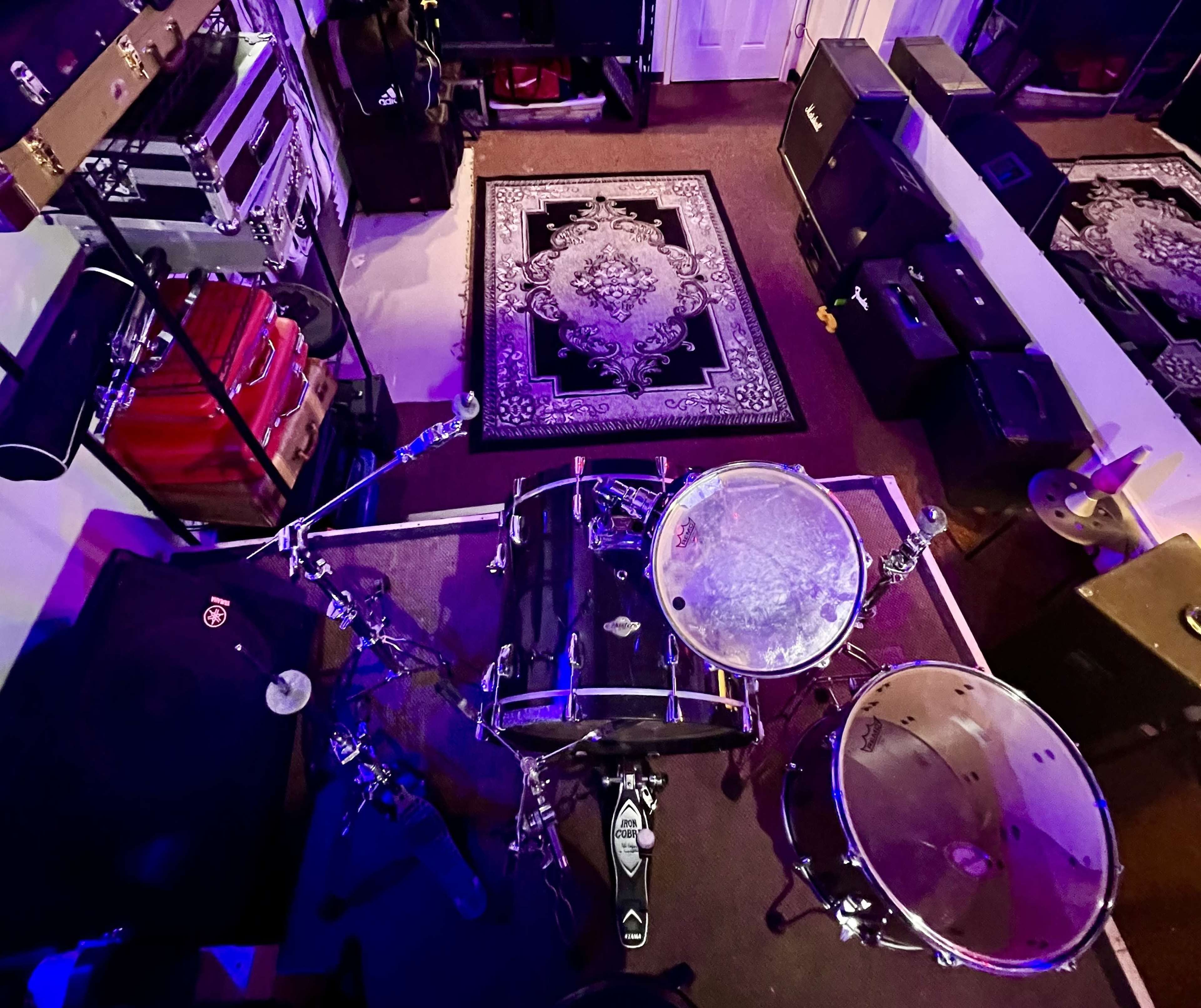 The image shows a drum set positioned on a carpet in a music studio filled with amplifiers and equipment.
