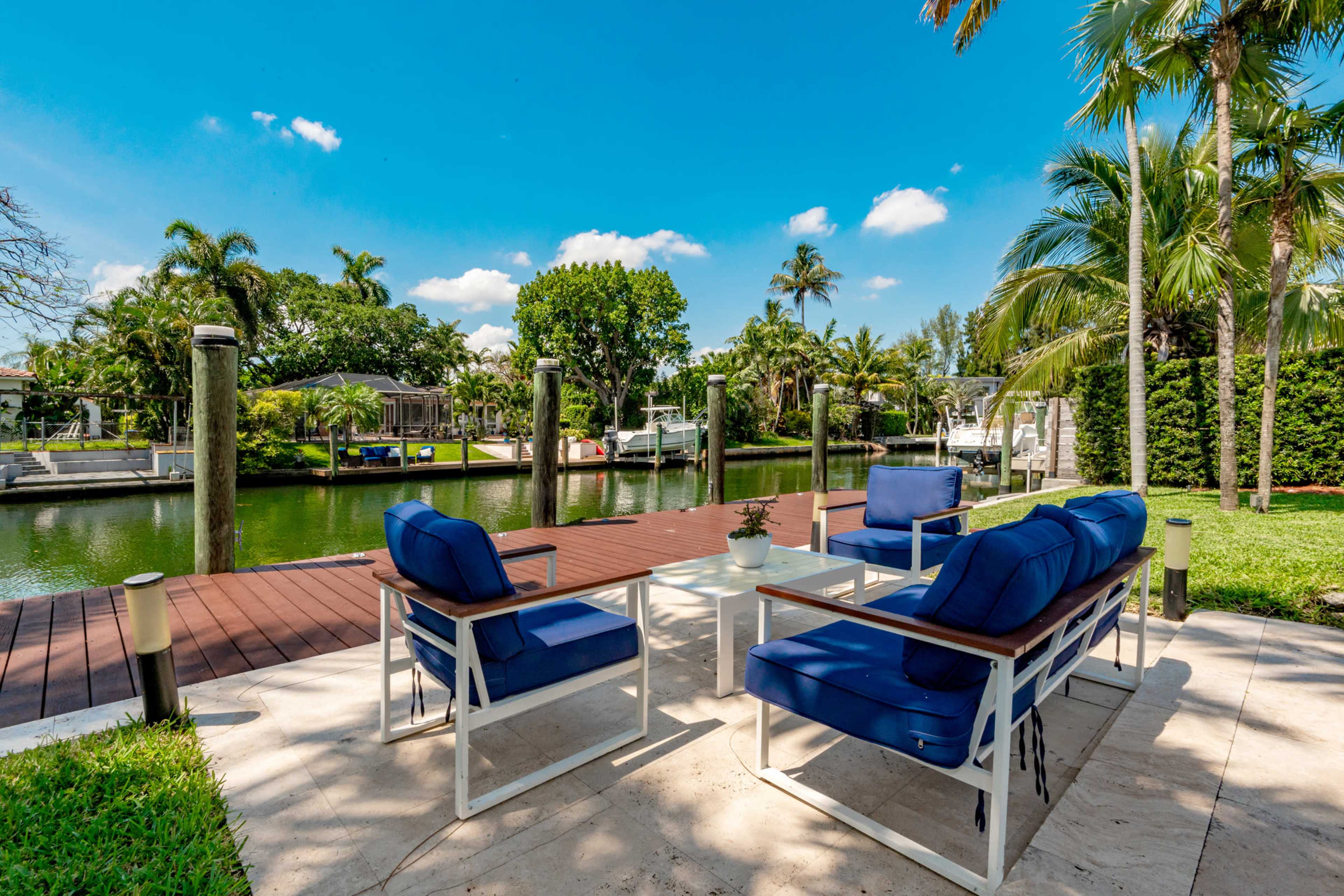 A patio with blue outdoor furniture overlooks a canal lined with palm trees and greenery under a clear blue sky.