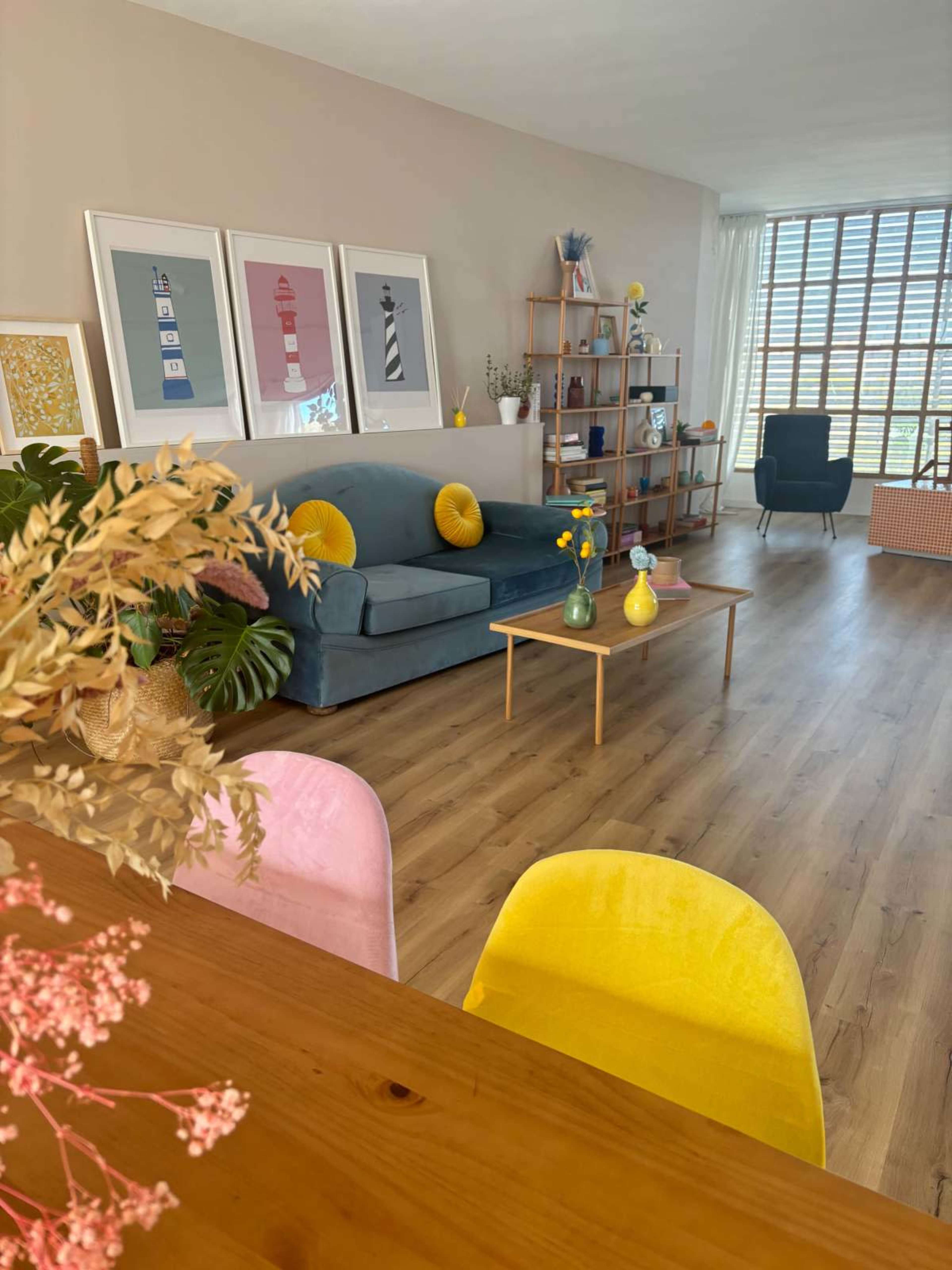 A brightly lit living room features a blue sofa with yellow cushions, a wooden coffee table, and artwork on the walls, with a dining area in the foreground.