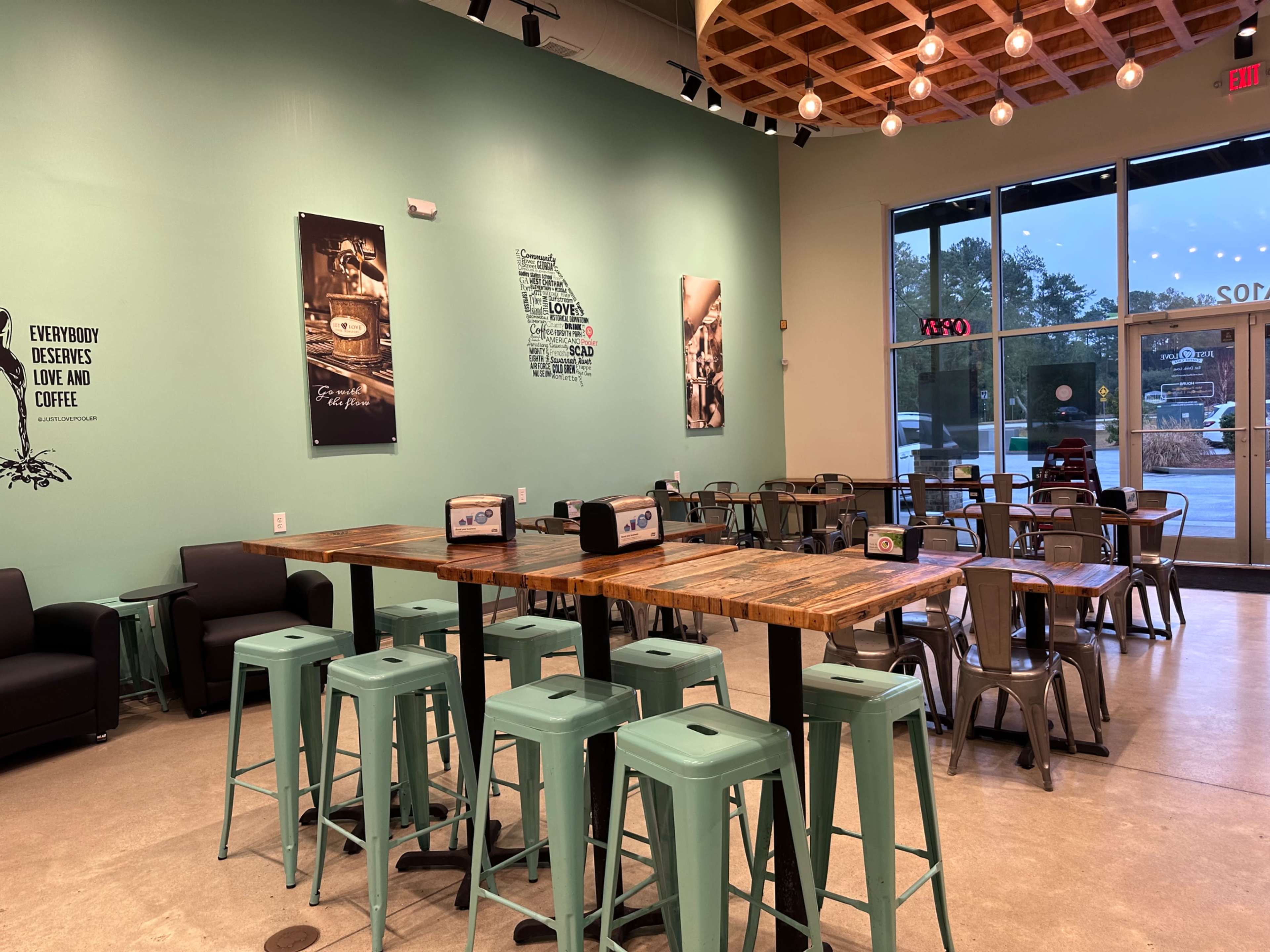 A cozy café interior with metal stools and wooden tables, featuring large windows and wall art.