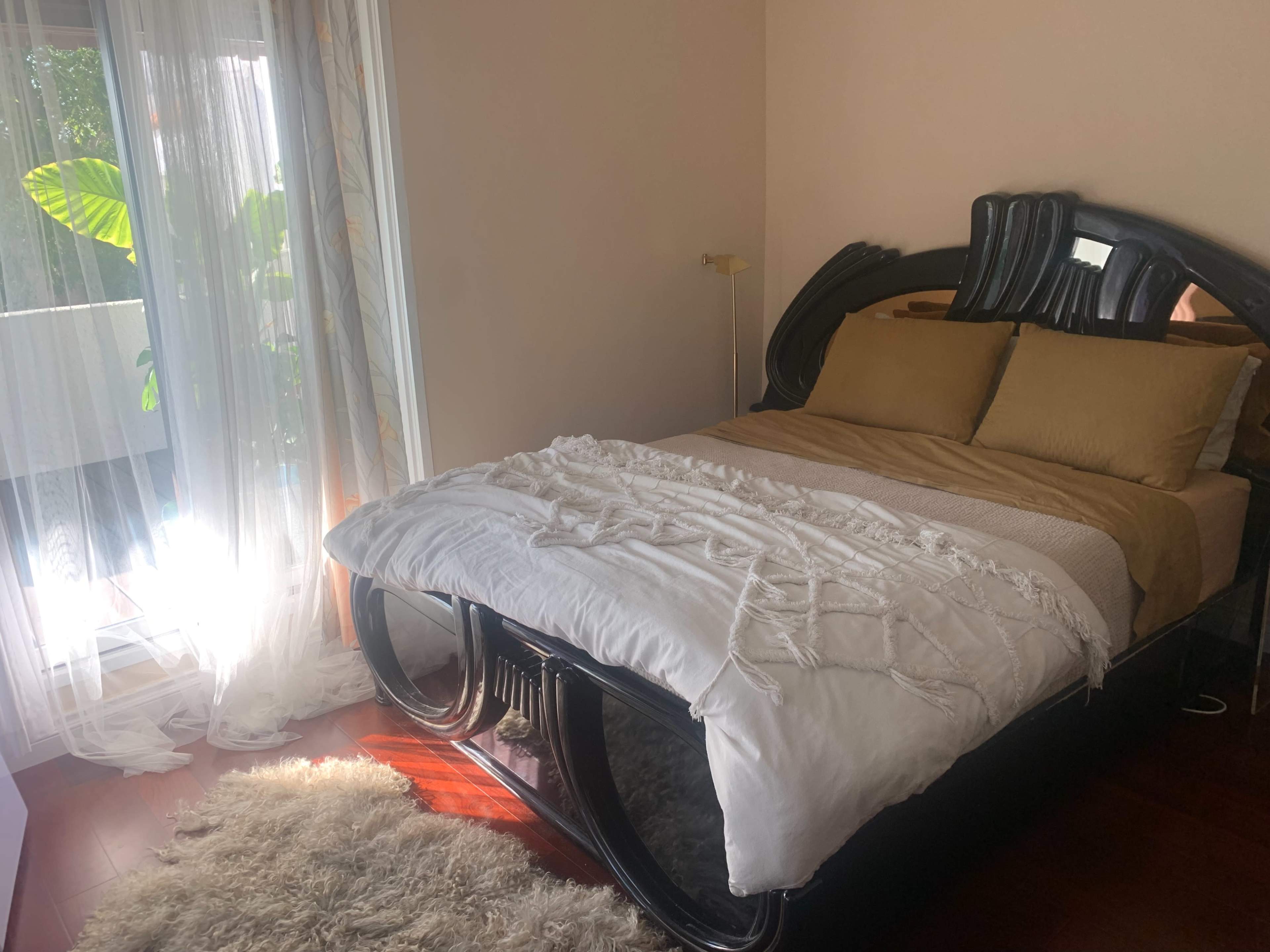 A bedroom features a black bed with a white bedspread and two golden pillows, illuminated by natural light from a nearby window.