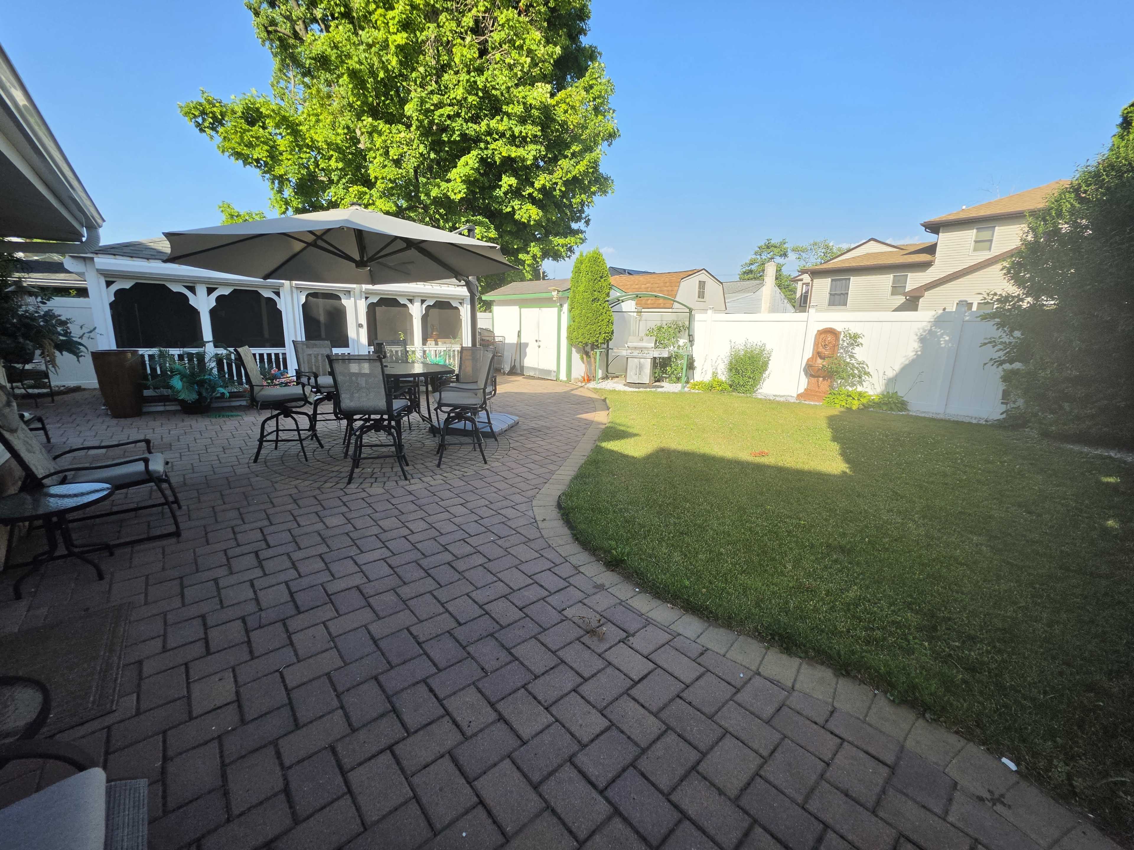 The image shows a backyard patio area with a stone pathway, patio furniture, a large umbrella, and a well-maintained lawn surrounded by a fence.