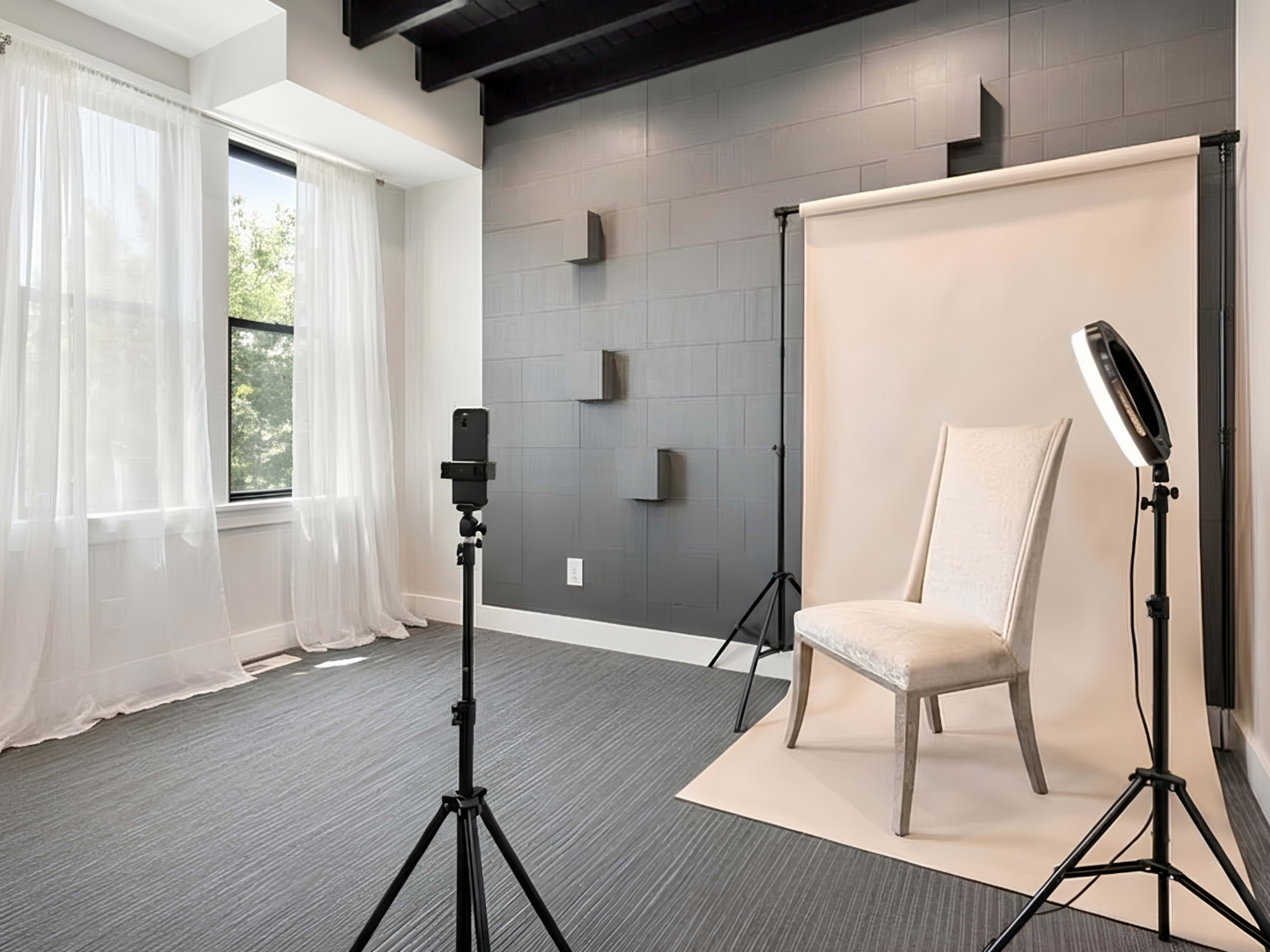 A simple photo studio features a single chair on a light backdrop, flanked by a camera on a tripod and a ring light, with large windows letting in natural light.
