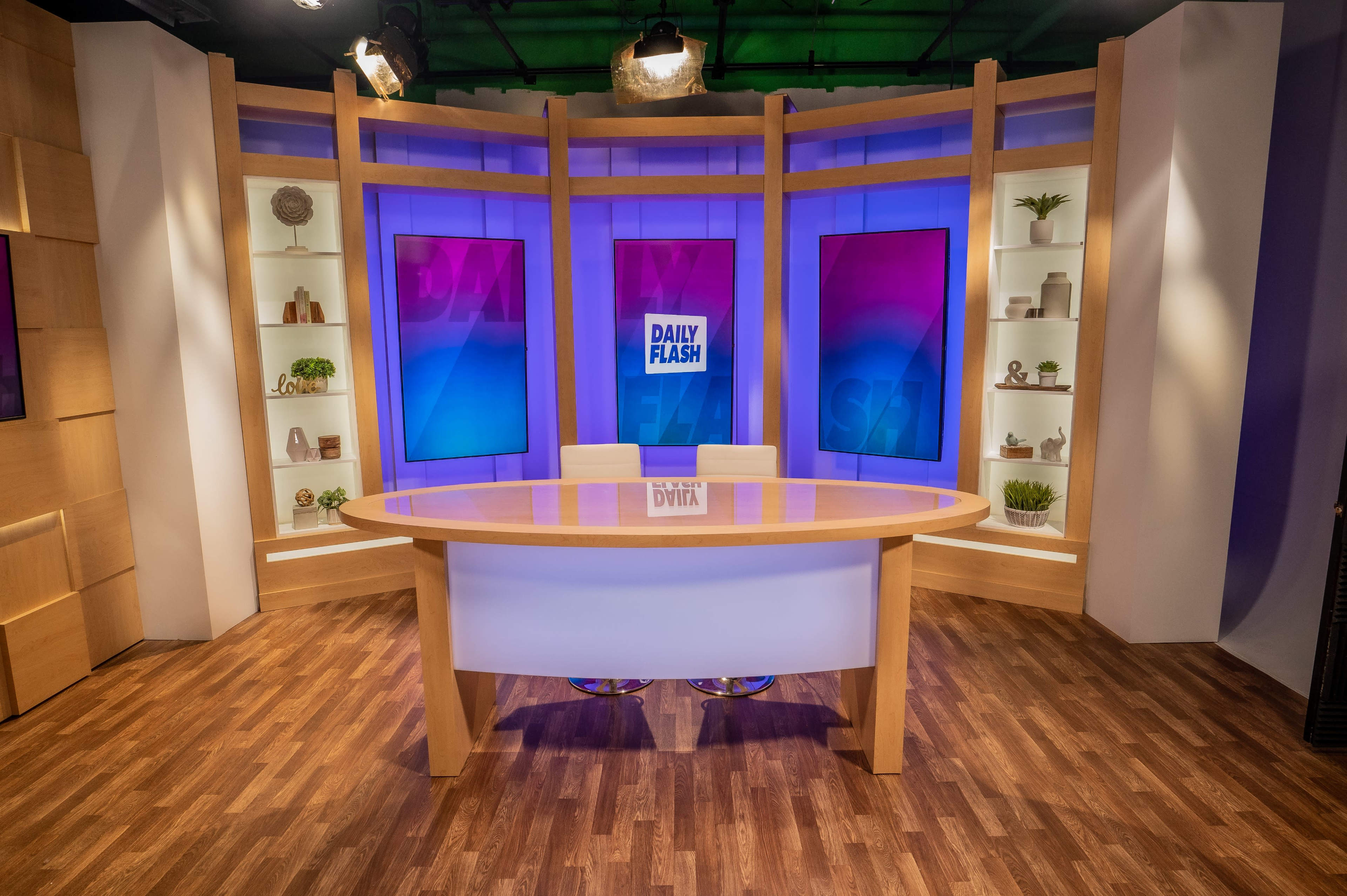 The image shows a modern television studio set with a round wooden table, flanked by blue and purple lit background screens displaying "DAILY FLASH."