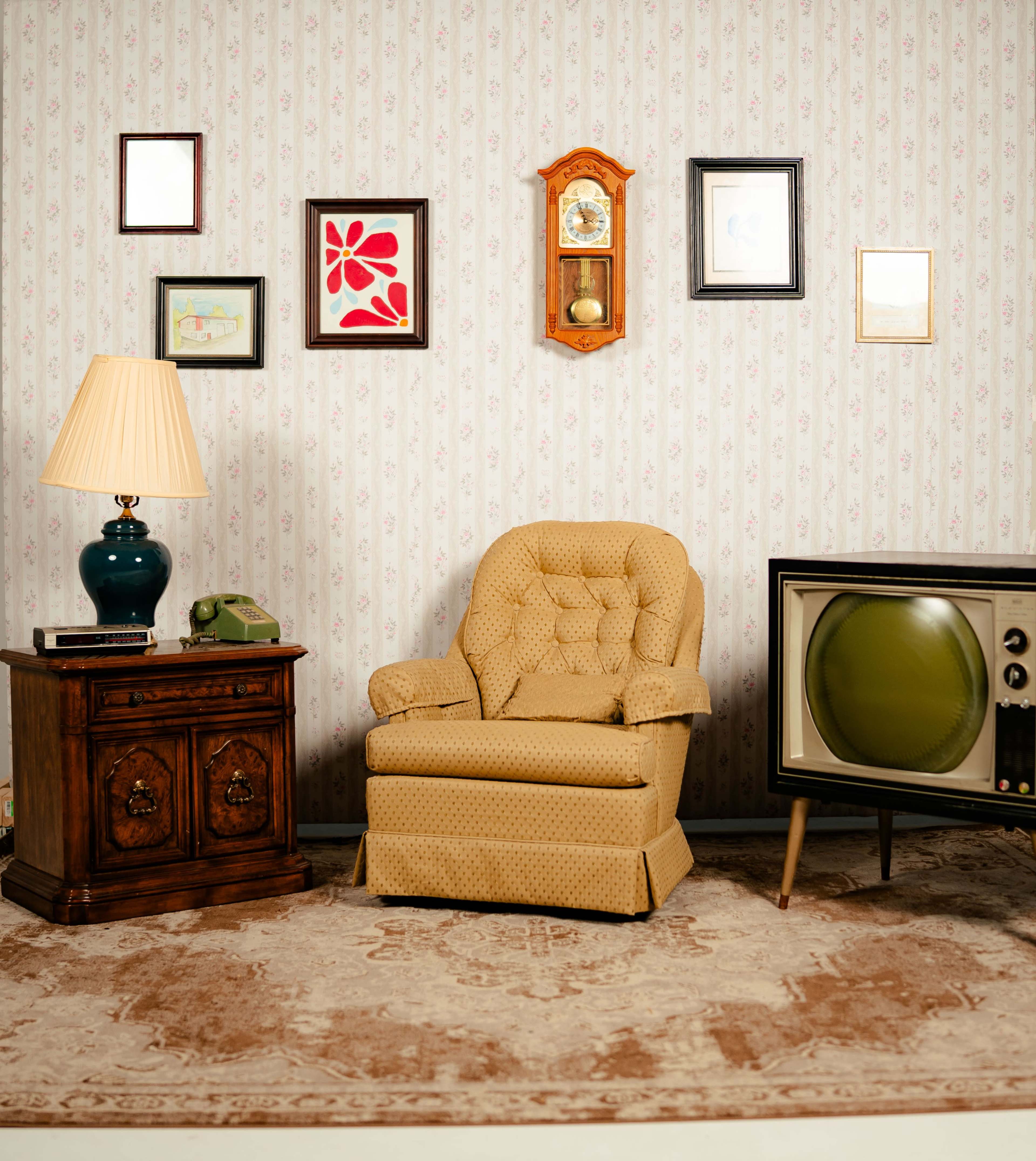 The image displays a vintage living room scene featuring a patterned armchair, a lamp, a wooden side table with a telephone, wall art, and an old television set.