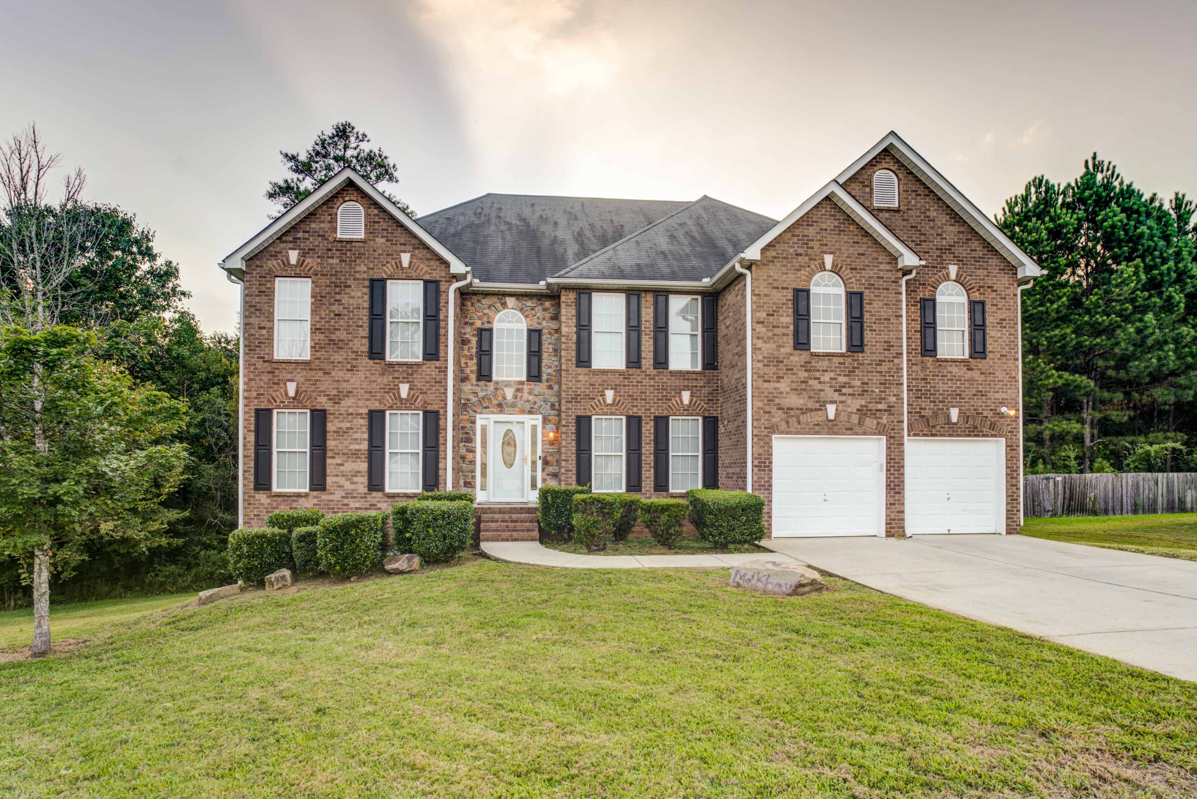 A two-story brick house with a symmetrical design, featuring a central front door, multiple windows, and an attached double garage, is situated on a lawn with trees in the background.