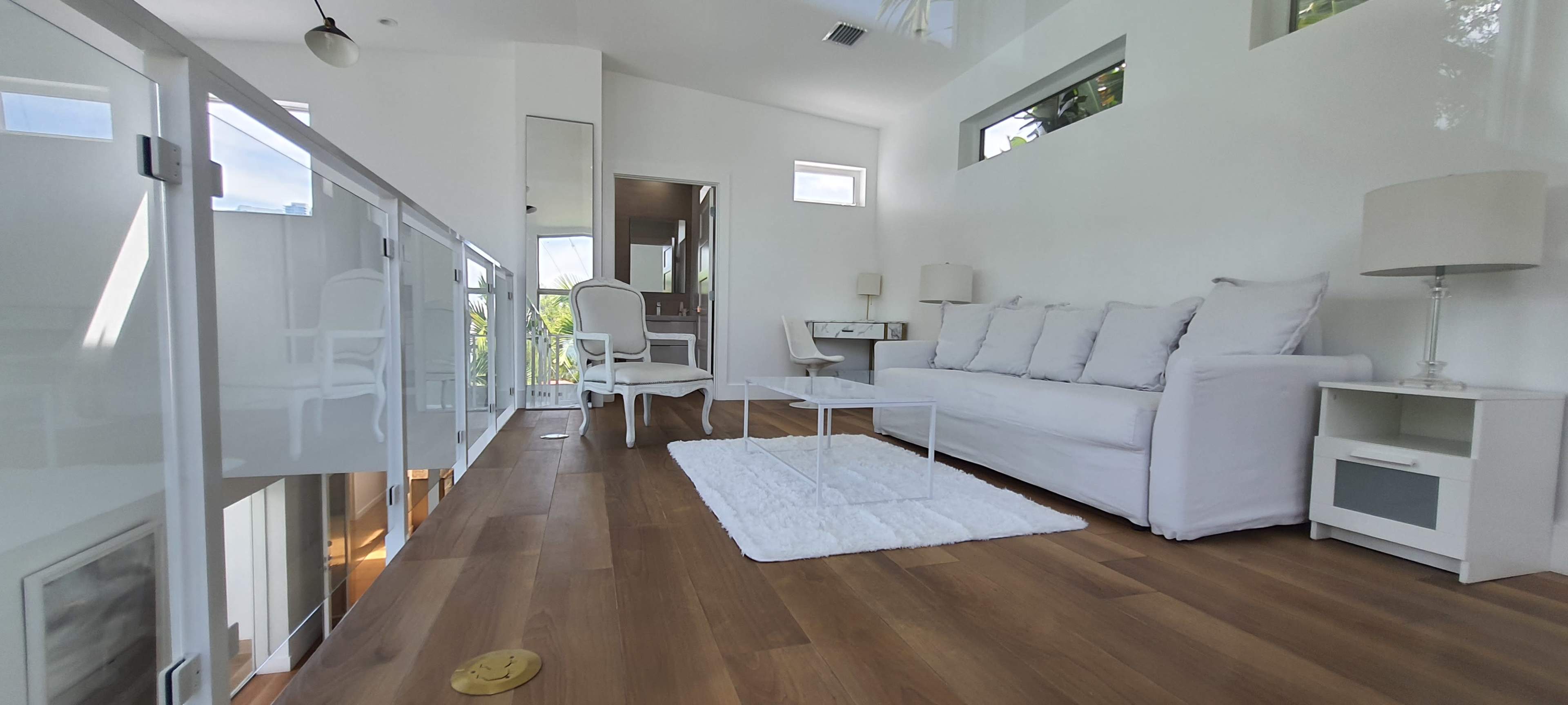 A bright, modern living space features a white sofa, a glass coffee table, and light wood flooring, with large windows allowing natural light to enter.
