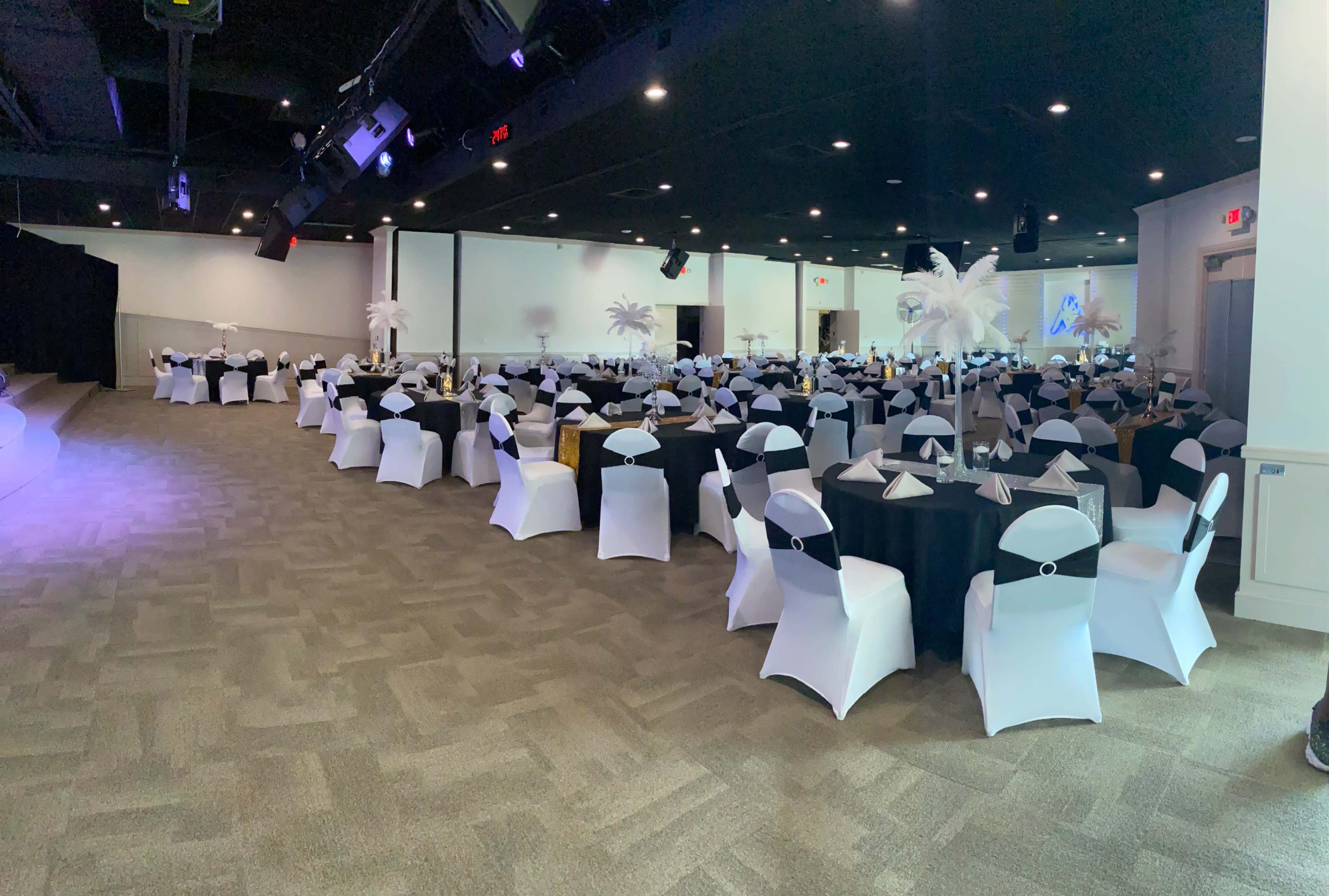 A spacious banquet hall is set up with round tables covered in black and white tablecloths, featuring neatly arranged place settings and decorative centerpieces.