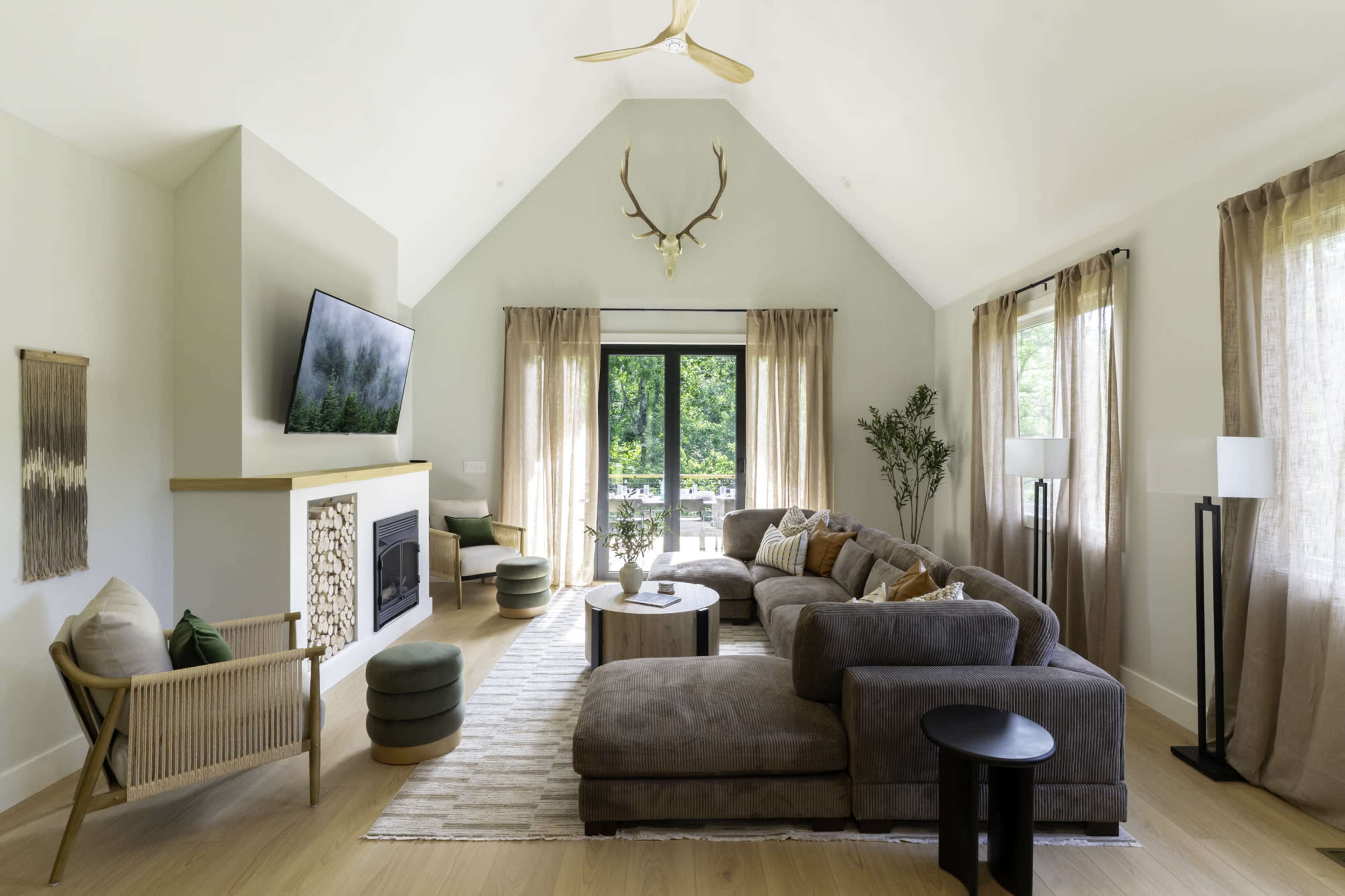 The image shows a modern living room with a large sectional sofa, a flat-screen TV mounted on the wall, large windows with sheer curtains, and a cozy decorative fireplace.