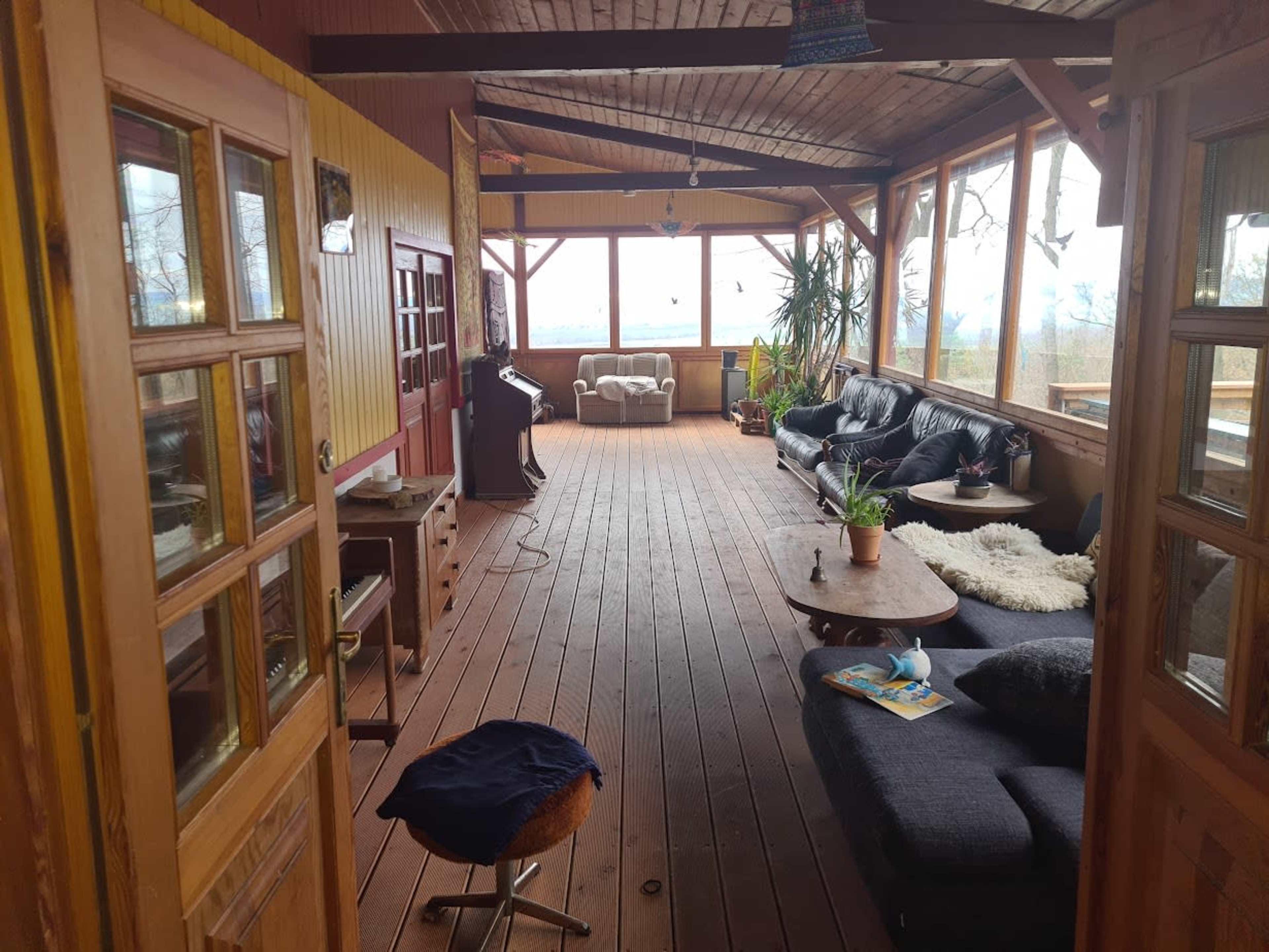 The image shows a spacious, enclosed porch with wooden walls, a wooden floor, and comfortable seating, illuminated by natural light from large windows.