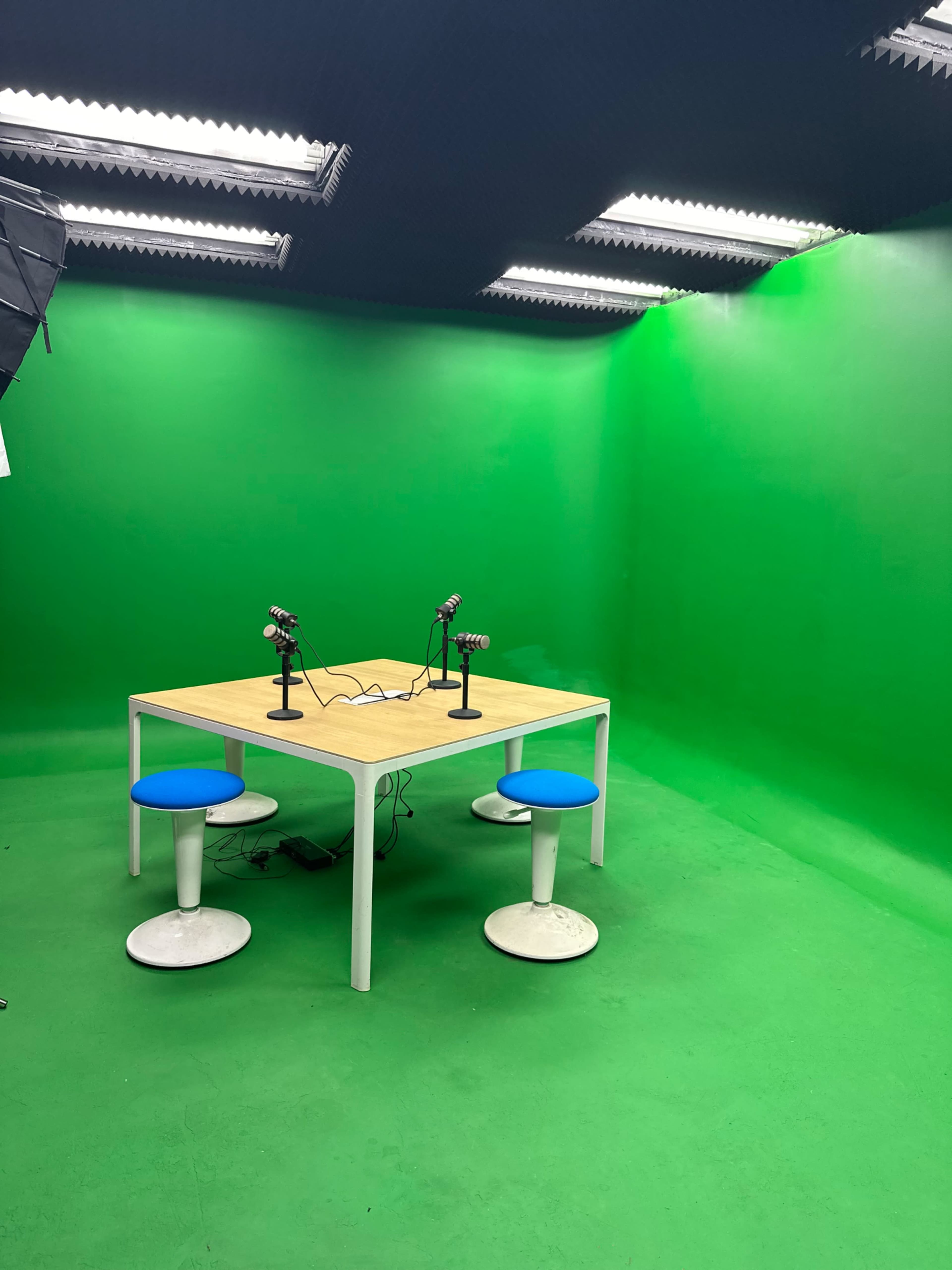 A green screen studio features a wooden table with two microphones and four stools.