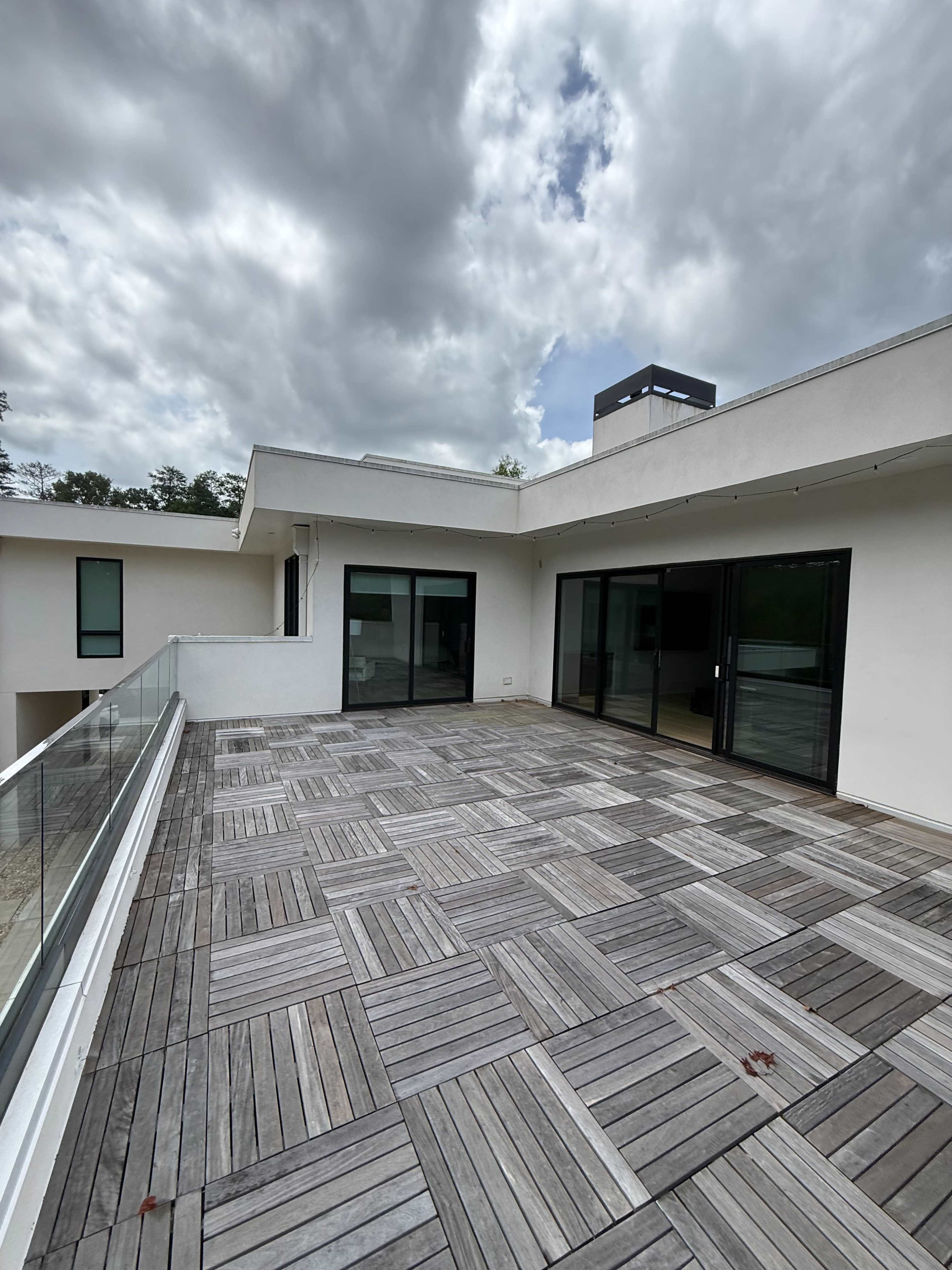 The image shows a spacious outdoor terrace with wooden flooring and large glass doors leading to an interior space, under a partly cloudy sky.