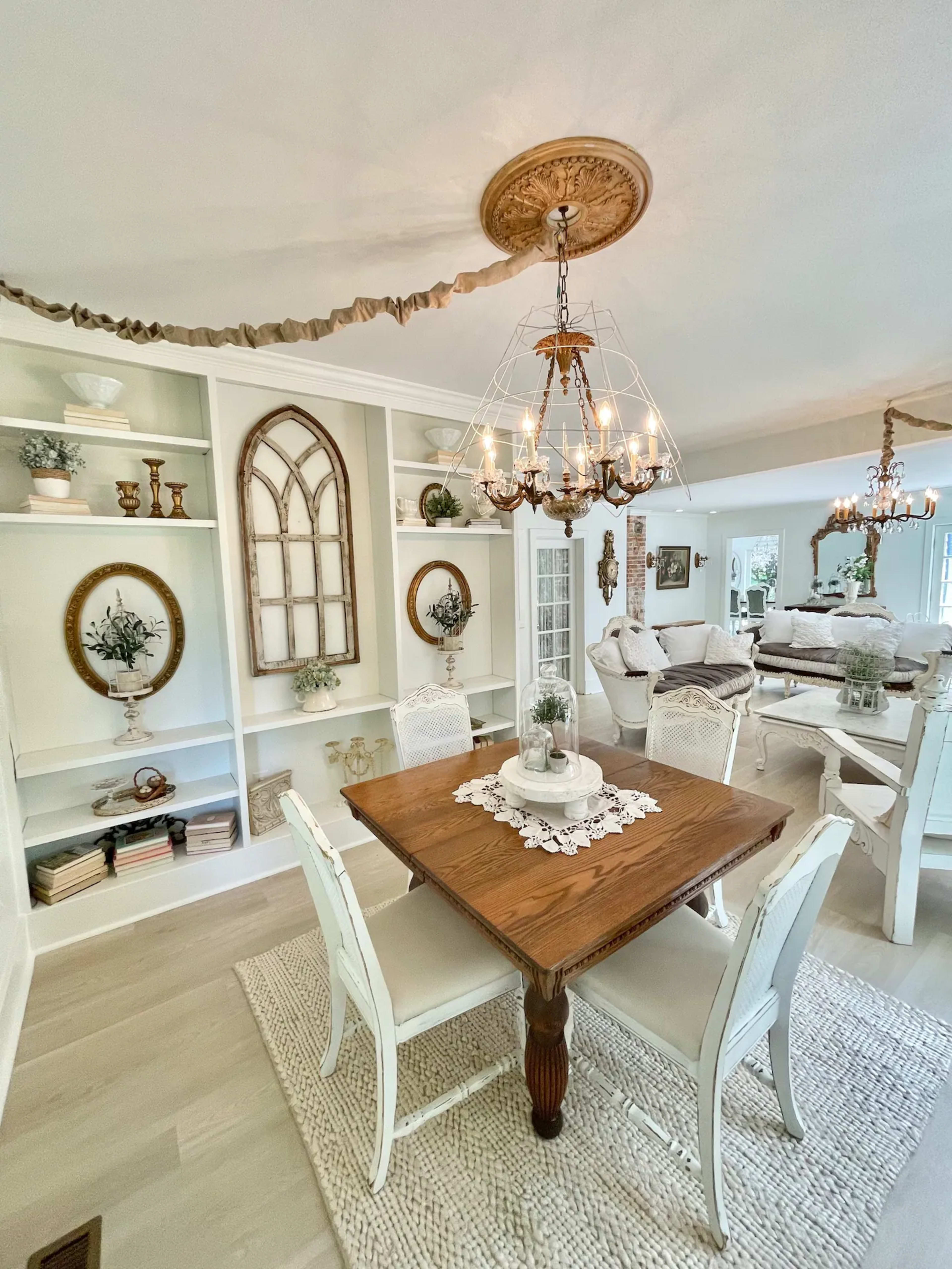 A wooden dining table surrounded by white chairs sits in a well-decorated dining area with built-in shelves and decorative elements.