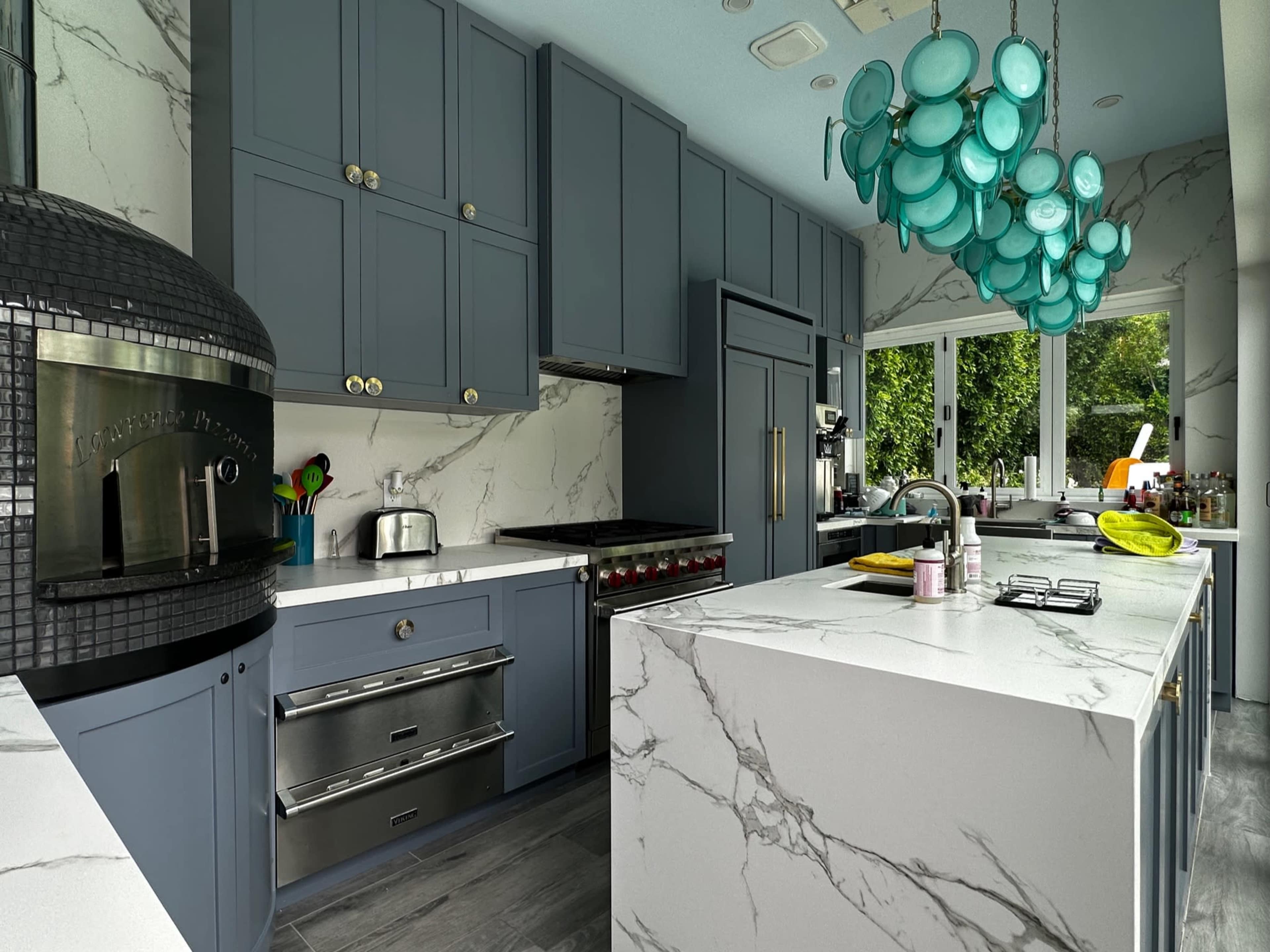 A modern kitchen with blue cabinets, a marble countertop, and a unique chandelier made of turquoise glass.