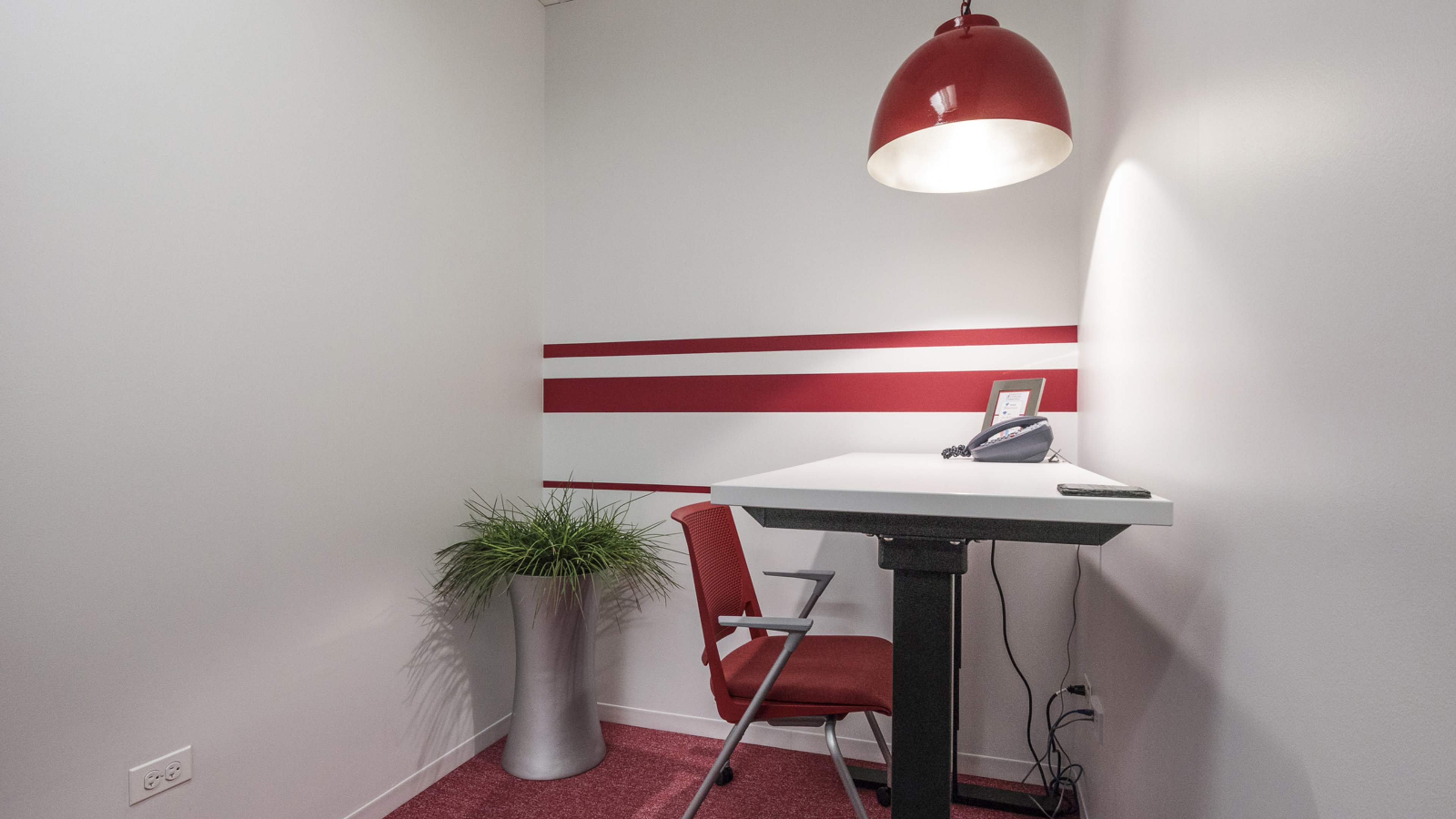A small office space features a red pendant light, a desk with a phone, a chair, and a potted plant against a white wall with red stripes.