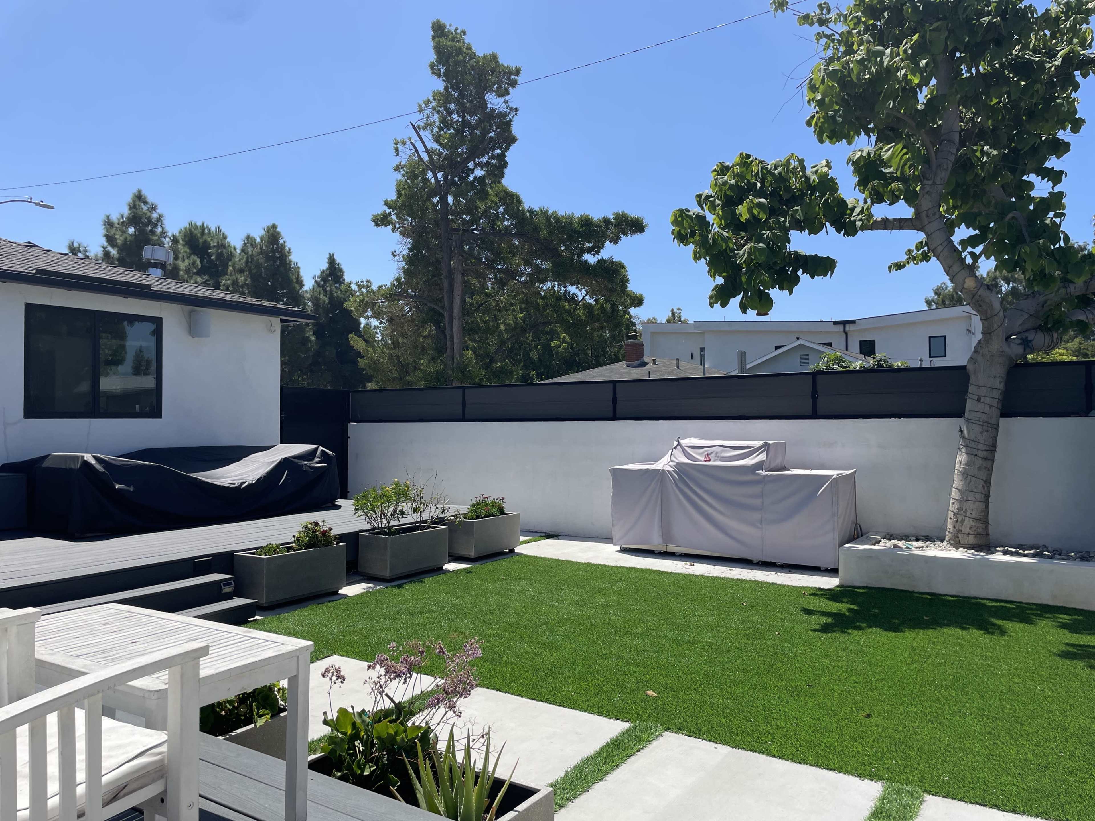 A backyard with artificial grass, planters, and outdoor furniture, featuring a covered barbecue grill and a blanketed outdoor unit against a backdrop of trees and blue sky.