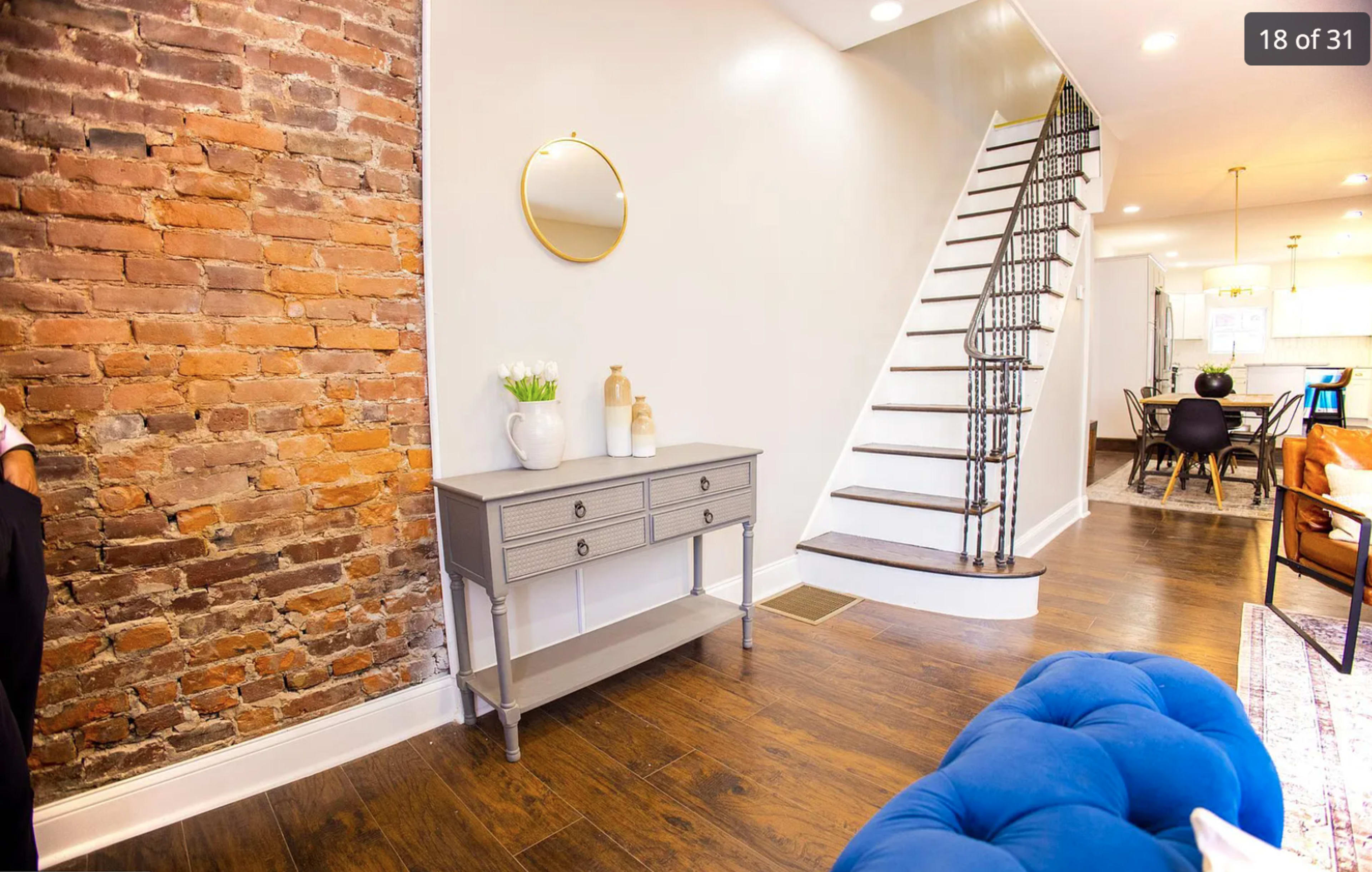 A hallway with a brick accent wall, a circular mirror above a gray console table, and a staircase leading to the upper floor.